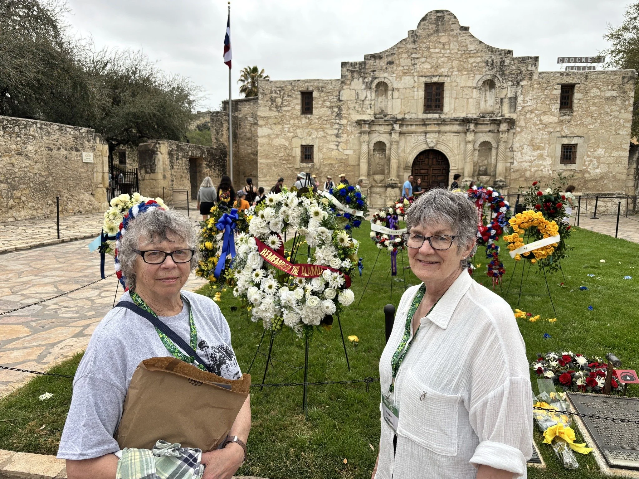 Two women standing outdoors in front of a historic stone building with a large floral display of white, red, yellow, and purple flowers. The woman on the left is holding a paper bag, and both are wearing glasses and tourist badges.