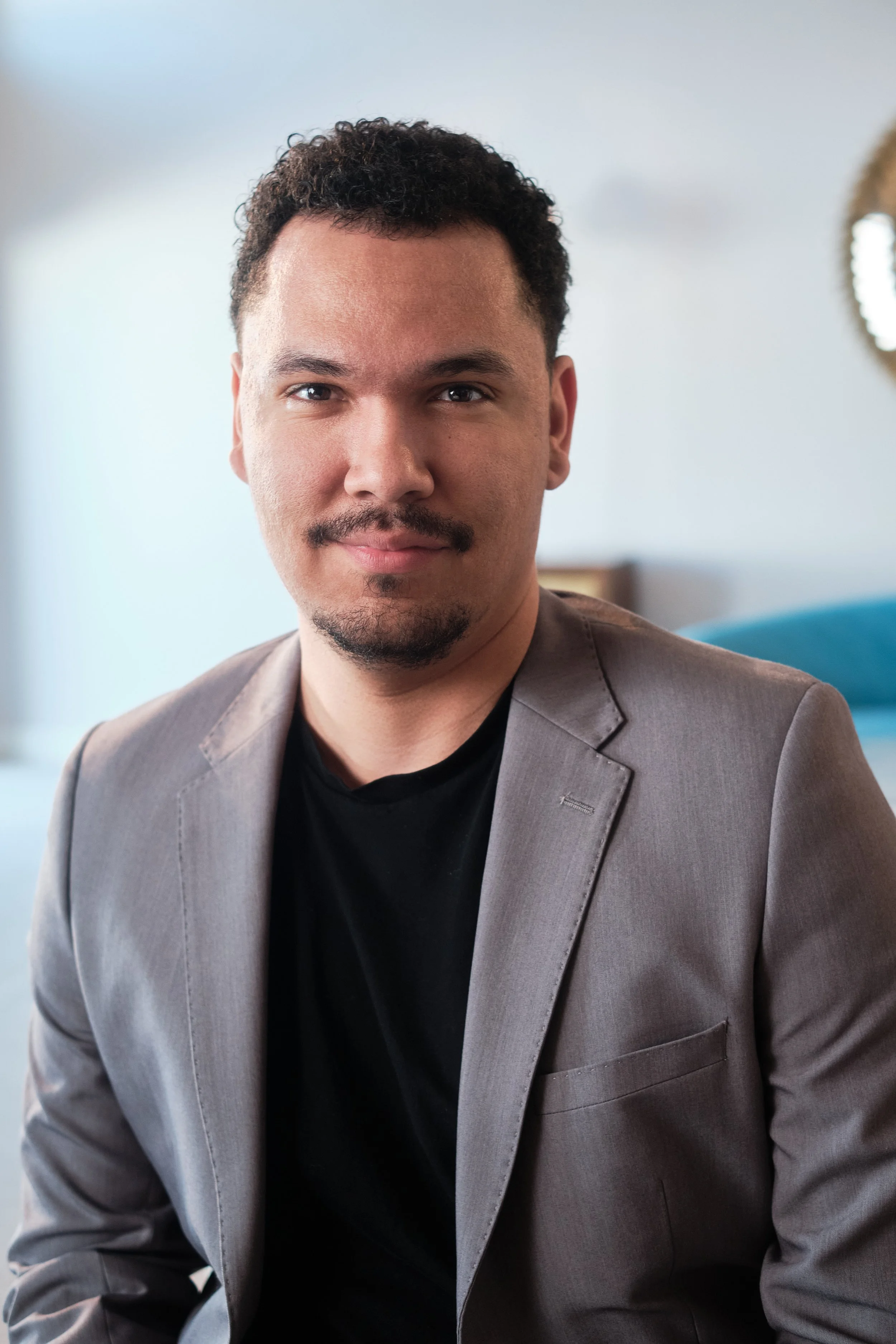 A man with short curly hair, a mustache, and a goatee, dressed in a light gray suit jacket over a black shirt, sitting indoors with a blurred background including a mirror and a blue object. Alex Johnson of Event Life TV Headshot Photograph