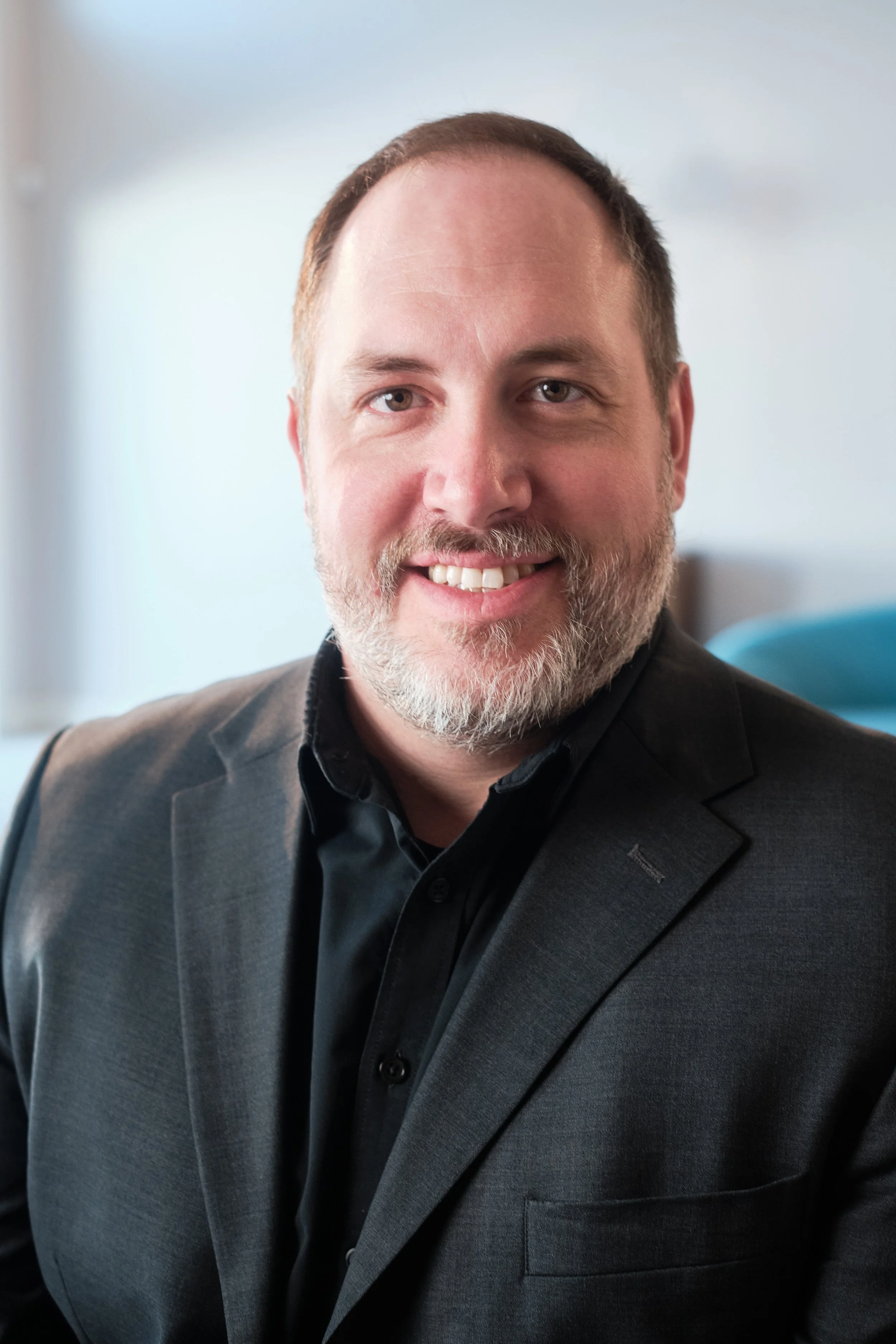 A man with a beard and short hair wearing a dark gray suit and black shirt, smiling, inside a bright room with a light-colored wall. Esteban Cuadros of Event Life TV Headshot photograph