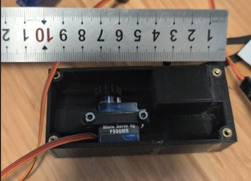 A black plastic servo motor housing with a small metal servo motor inside, connected to wires, and a metal ruler measuring approximately 15 centimeters or 6 inches in length placed on top.