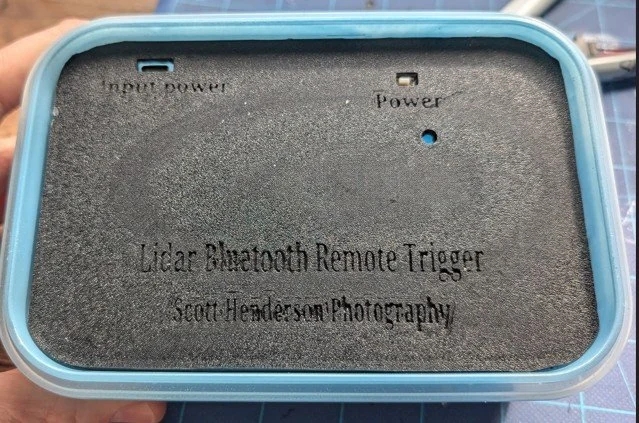 A blue rectangular box with a foam interior labeled 'Lidar Bluetooth Remote Trigger Scott Henderson Photography.' The interior has two small metal contact points labeled 'input power' and 'power.'
