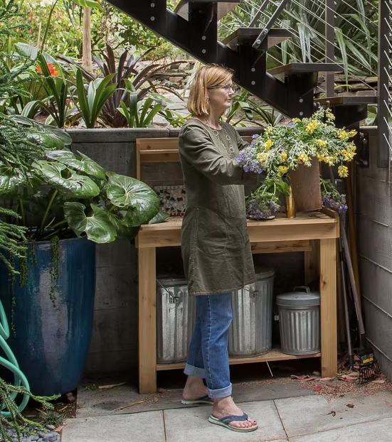 A woman with glasses and blonde hair in an outdoor garden area, holding a bouquet of yellow and purple flowers, standing next to a wooden table with large green plants and gardening supplies around her.
