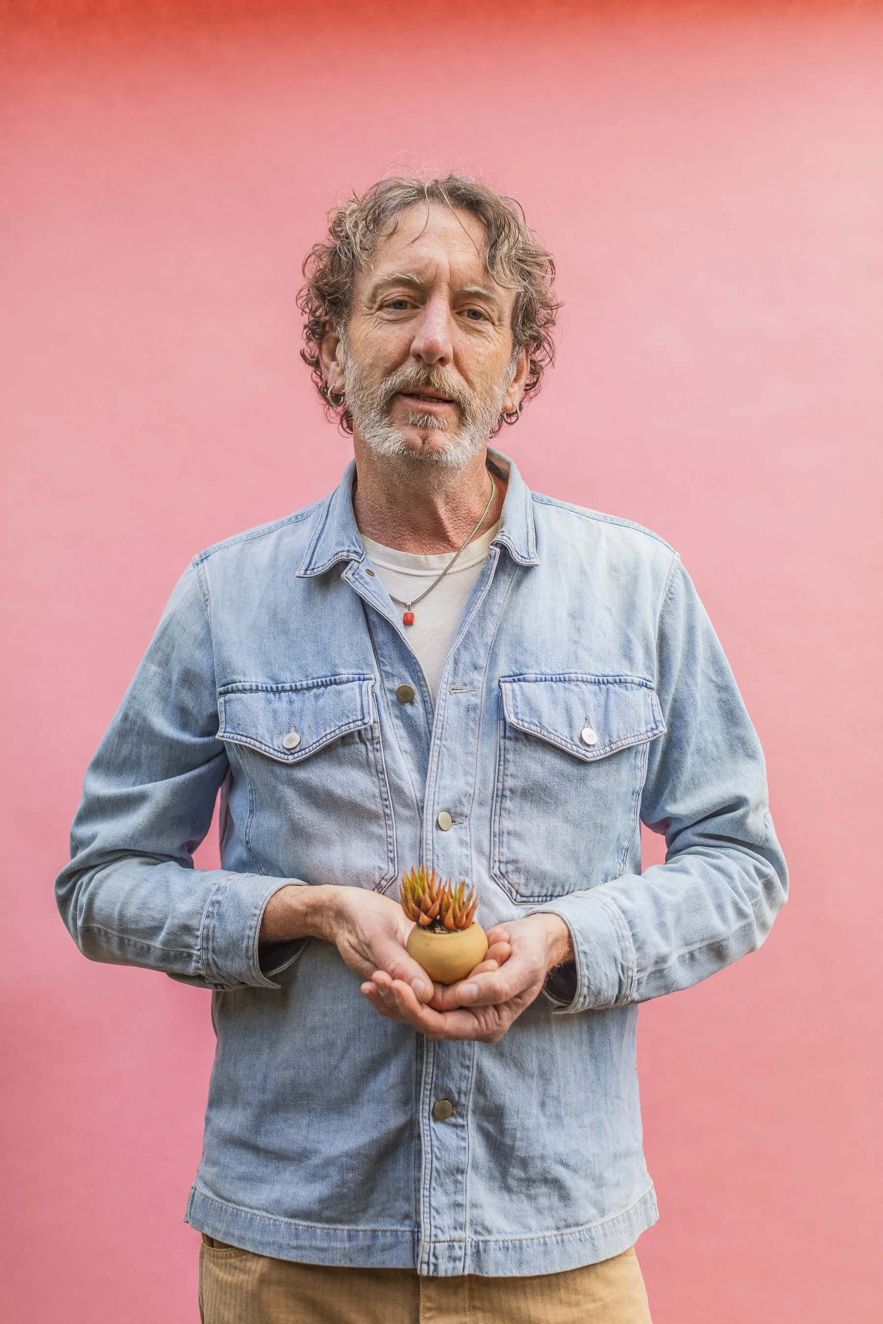 Clarke de Mornay, longtime staff member at Flora Grubb Gardens, holding a small clay pot with succulents, standing against a pink background.