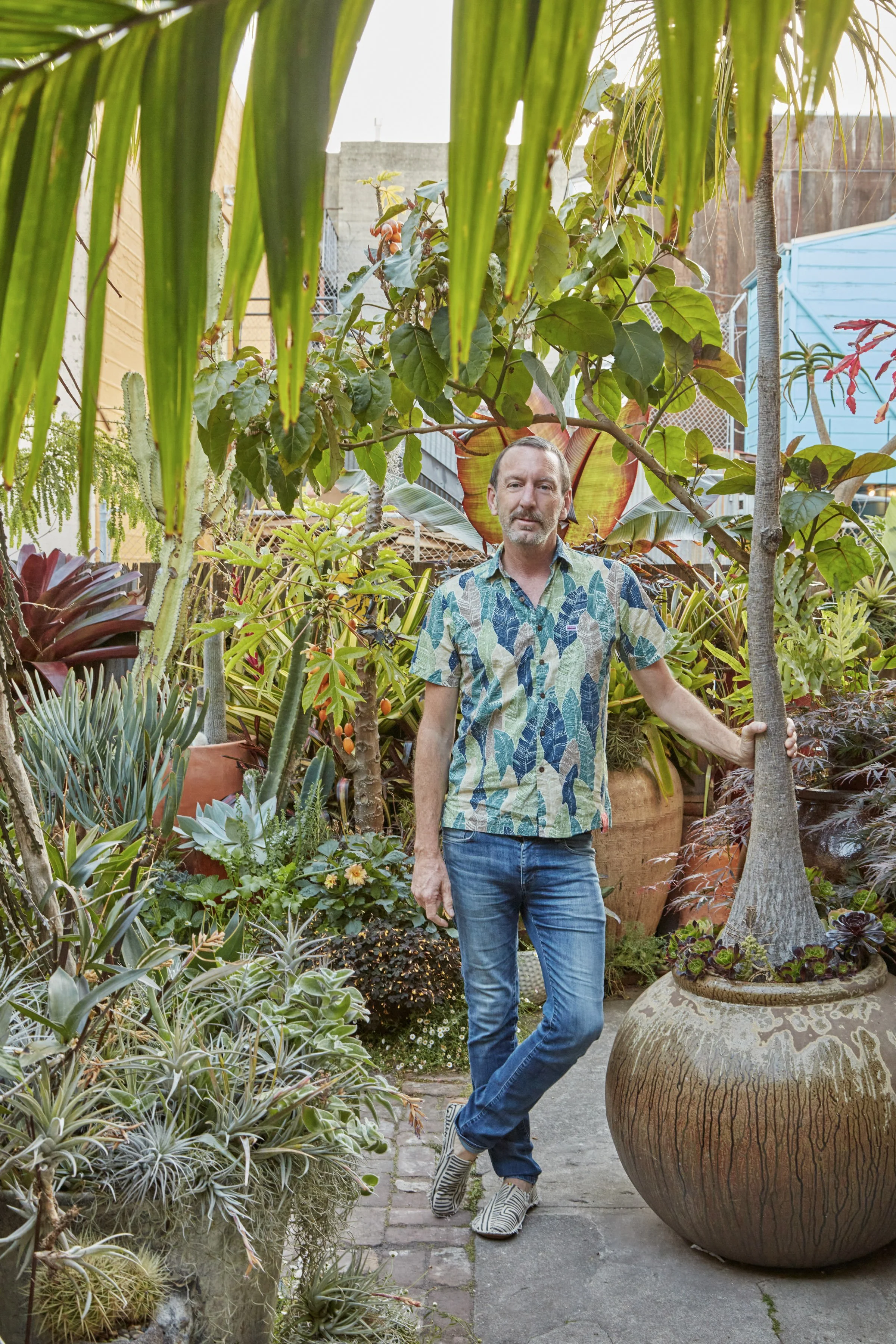 A man in a colorful, leaf-patterned shirt and jeans stands in a lush outdoor garden surrounded by various potted plants and greenery.