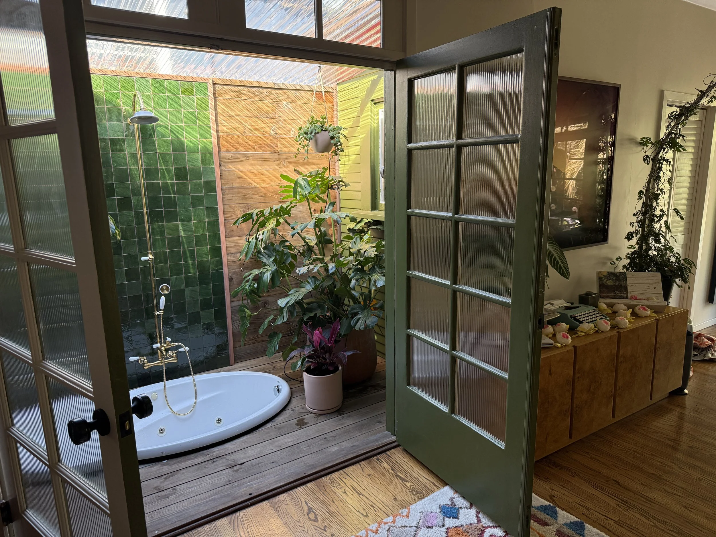 A bathroom greenhouse with an outdoor bath tub and shower space that has green tile walls, wooden floor, and a ceiling made of transparent corrugated material. The outdoor space contains potted plants. Designed with Terri Loenthal by Clarke De Mornay