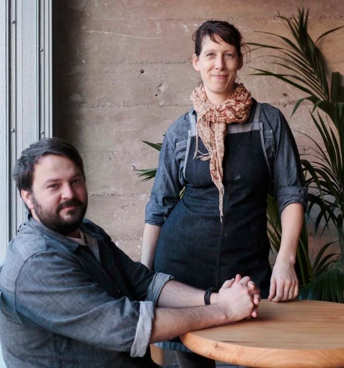 A woman standing next to a man seated at a wooden table in an indoor setting with a concrete wall and green plants in the background.