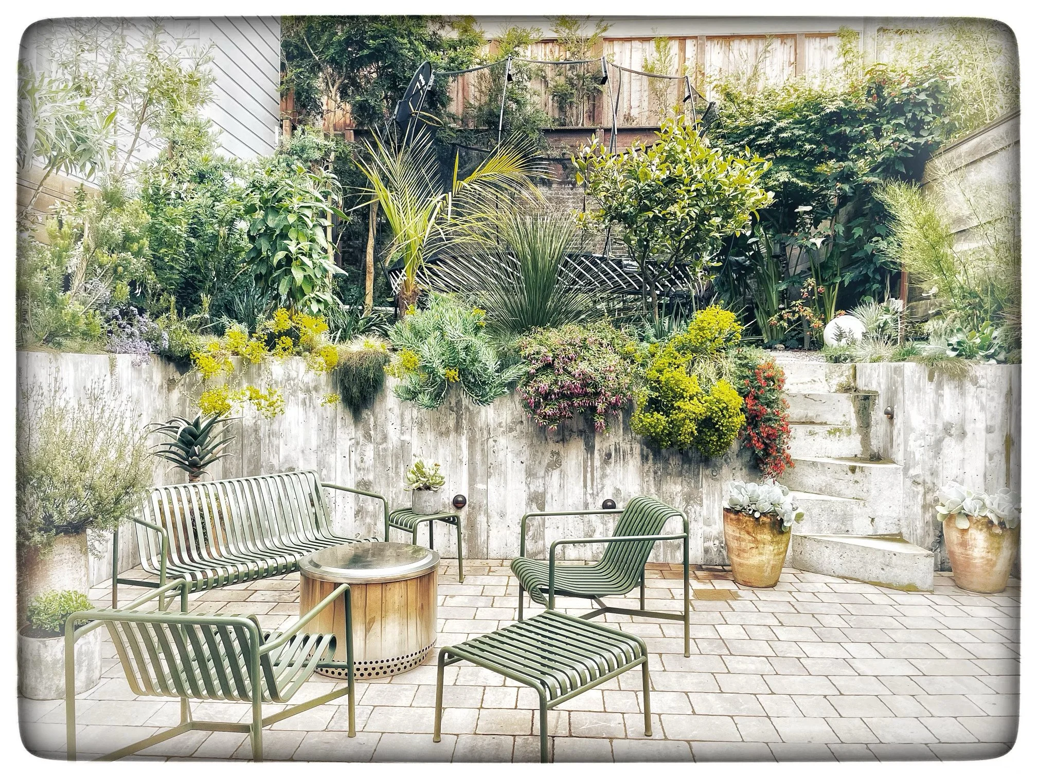 A cozy outdoor patio space with trees, plants, and flowers on a concrete and brick patio, including a round fire pit surrounded by metal chairs with striped cushions.