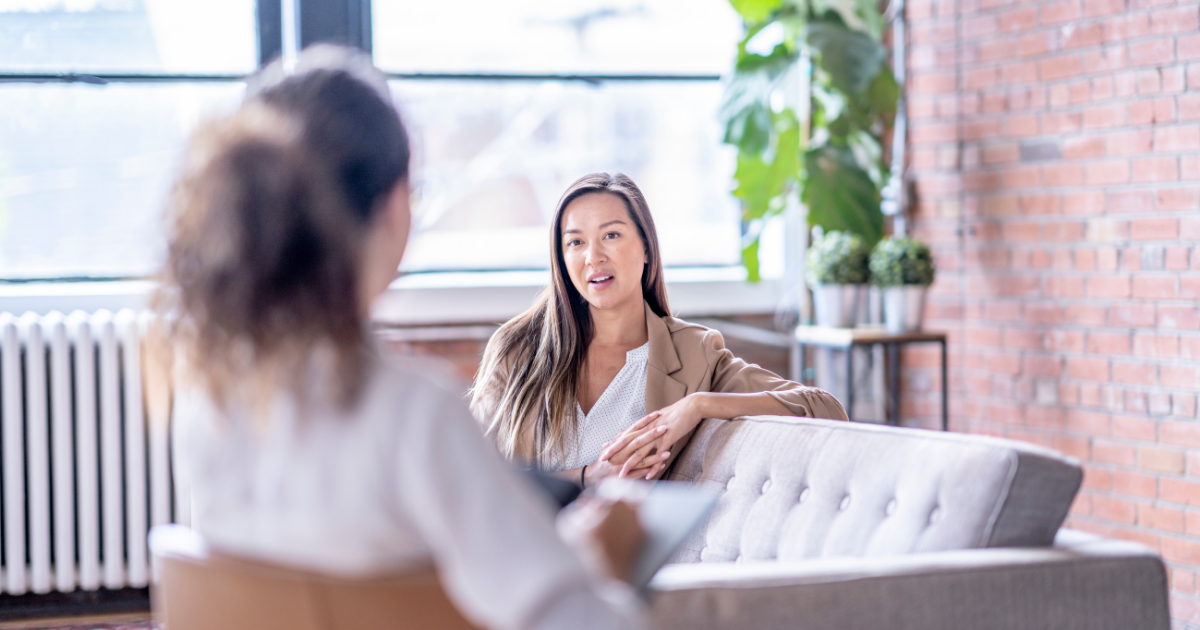 Two women having a conversation in a bright, modern office with large windows, a brick wall, and indoor plants.