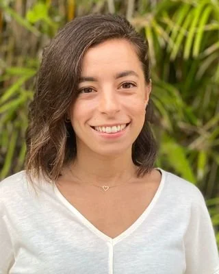 A woman smiling outdoors with green foliage in the background.