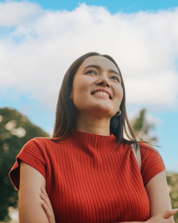 A woman with long dark hair wearing a red ribbed short-sleeve shirt, smiling and looking up outdoors with a blue sky and trees in the background.