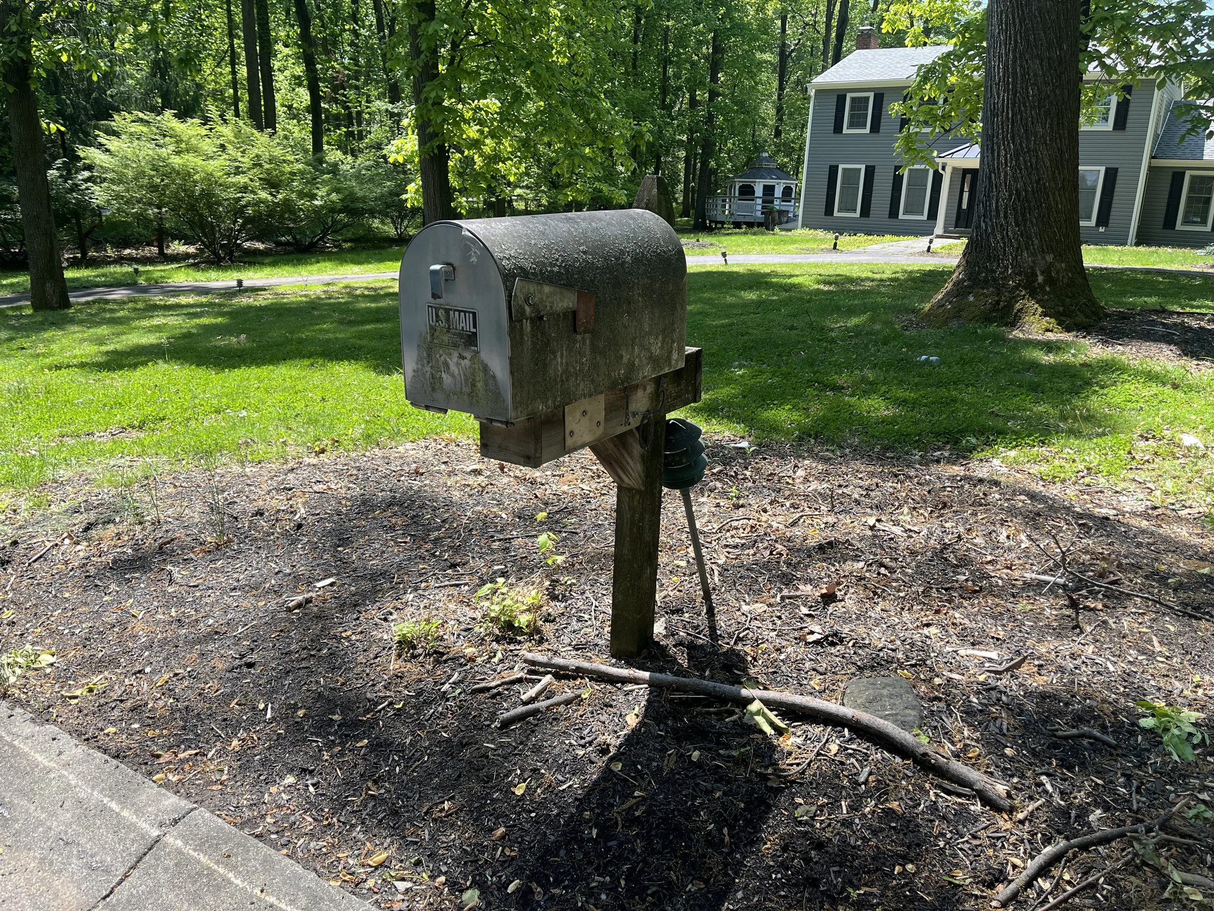 A mailbox with a rounded top and a U.S. Mail sticker, mounted on a wooden post, located on a grassy area near a driveway with a house in the background. The scene is shaded by trees in a suburban setting.