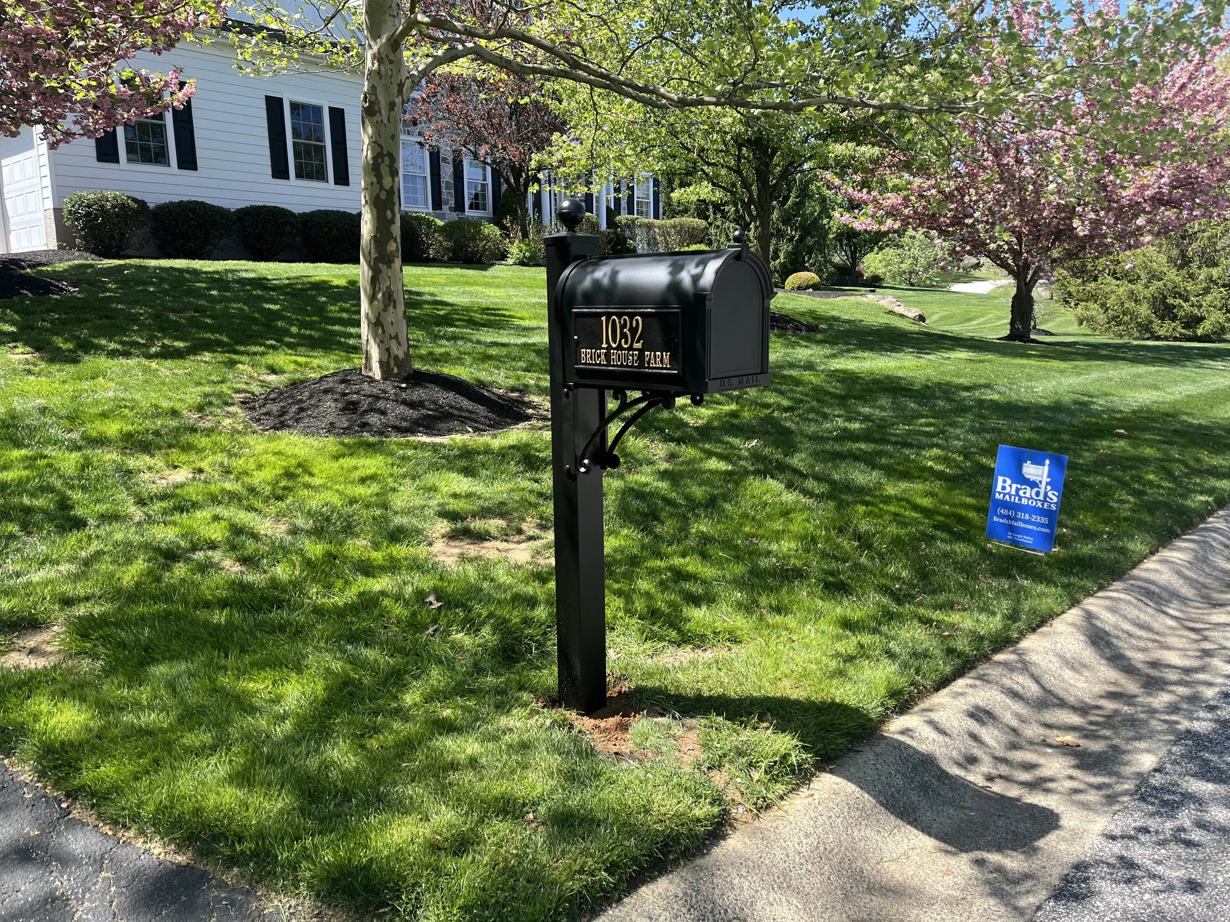 A black mailbox with gold numbers reading 1032 and the text 'Brick House Farm' is positioned on a lawn in front of a house, surrounded by blooming pink and purple flowering trees and lush green grass.