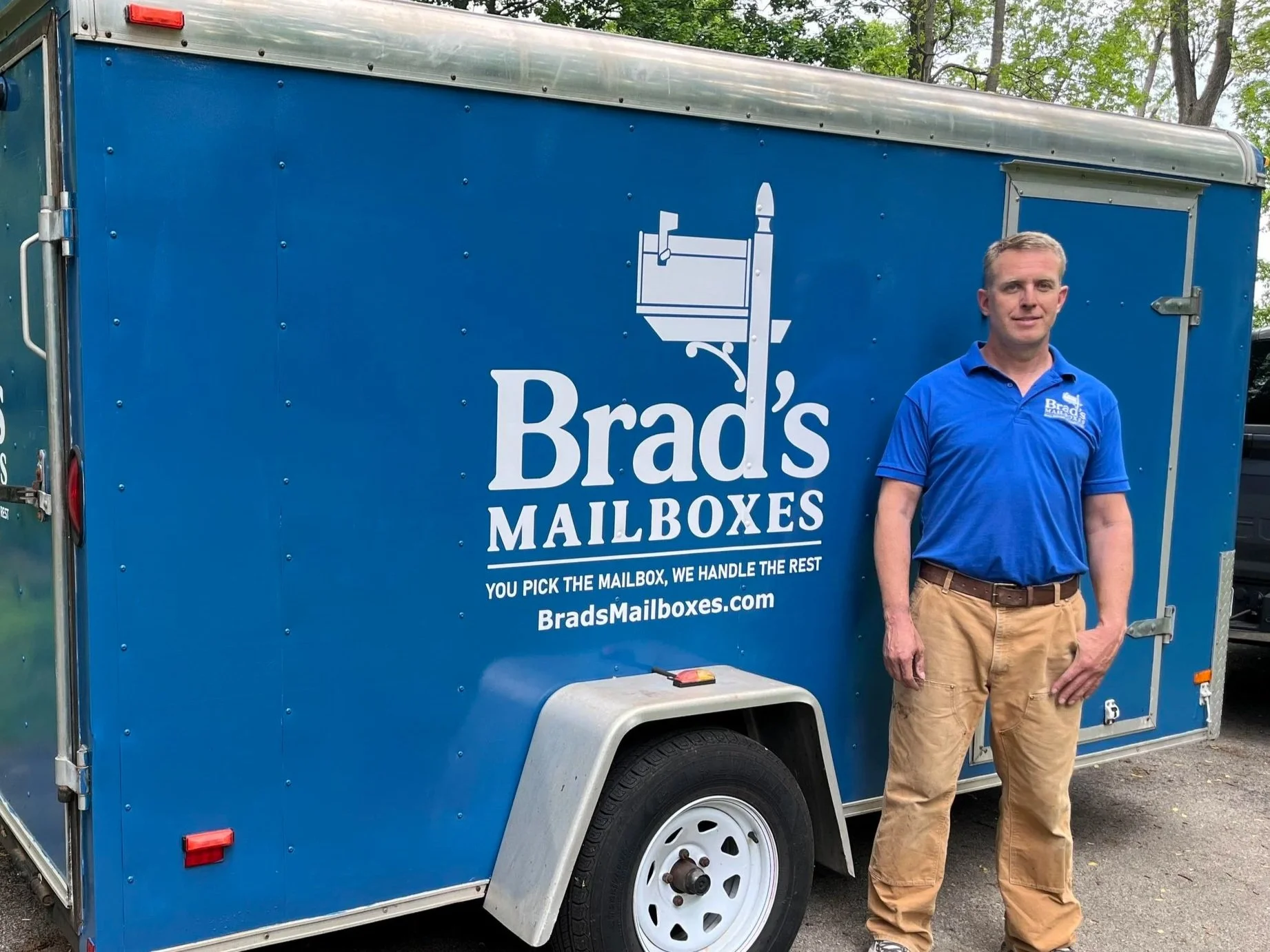 A man standing next to a blue mailbox delivery trailer with the name 'Brad's Mailboxes' printed on the side. He is wearing a blue polo shirt and khaki pants, with trees in the background.