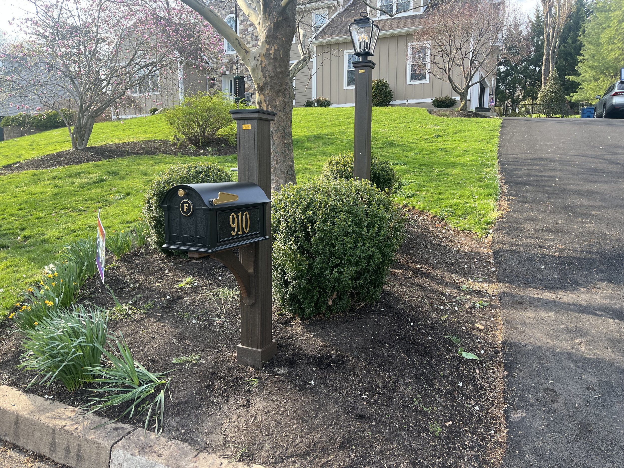 A front yard with green grass, small bushes, and blooming trees in front of a beige house. A black mailbox with the house number 910 is in the foreground. Two gray posts hold a porch light and a flag holder, with a driveway to the right leading toward the house and a parked car at the far end.