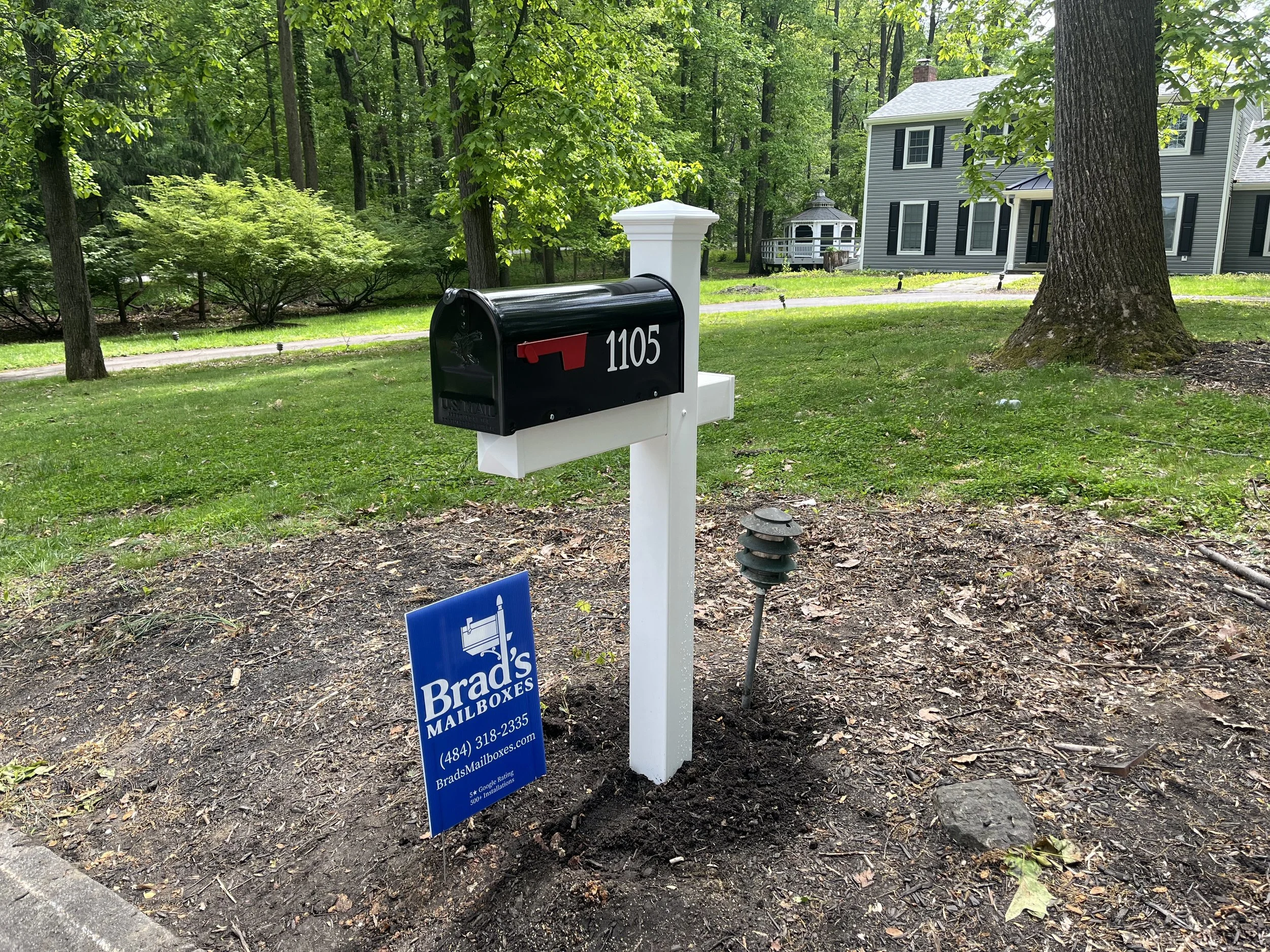 A black mailbox with the address 1105, mounted on a white post, is surrounded by a grassy yard with trees and bushes. A blue sign with white text advertising Brad's Mailboxes is placed on the ground nearby, and a house with dark shutters and a porch is visible in the background amidst a wooded area.