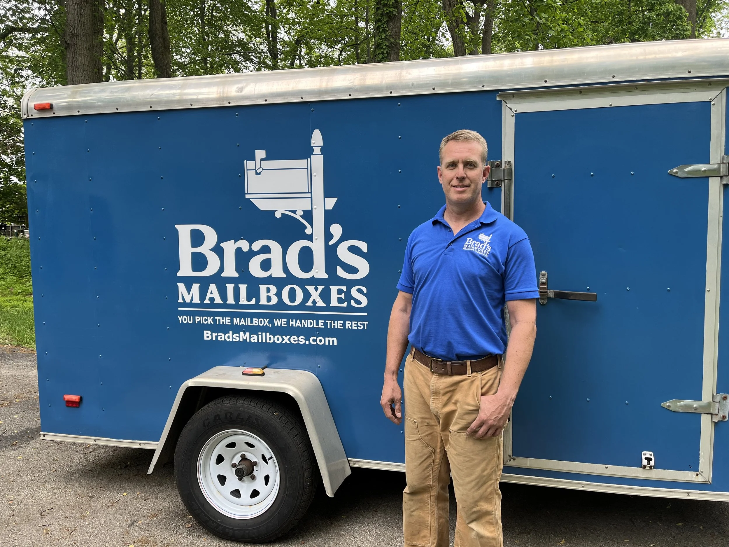 Man standing beside a blue mailbox trailer with 'Brads Mailboxes' logo, wearing a blue polo shirt and khaki pants, outdoors with trees in the background.