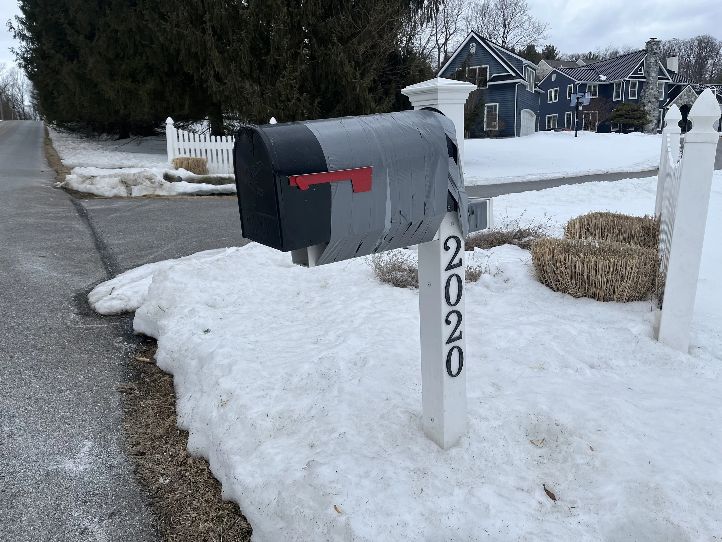 A damaged mailbox with duct tape holding it onto the post, with snow in the background