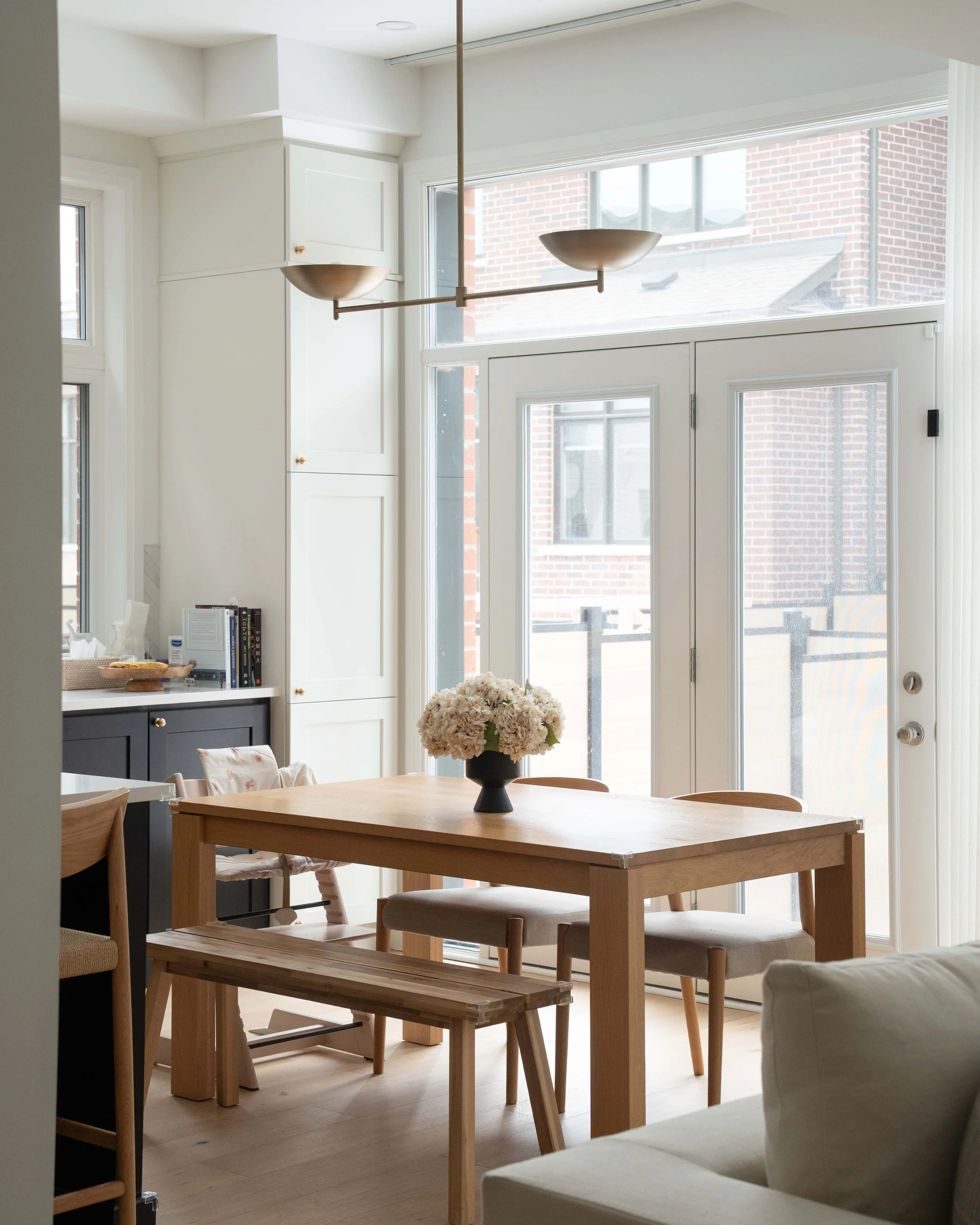 A dining area with a wooden table and chairs, a black vase with white flowers, large windows revealing a brick building outside, a white wall with built-in storage, and a modern light fixture hanging from the ceiling.