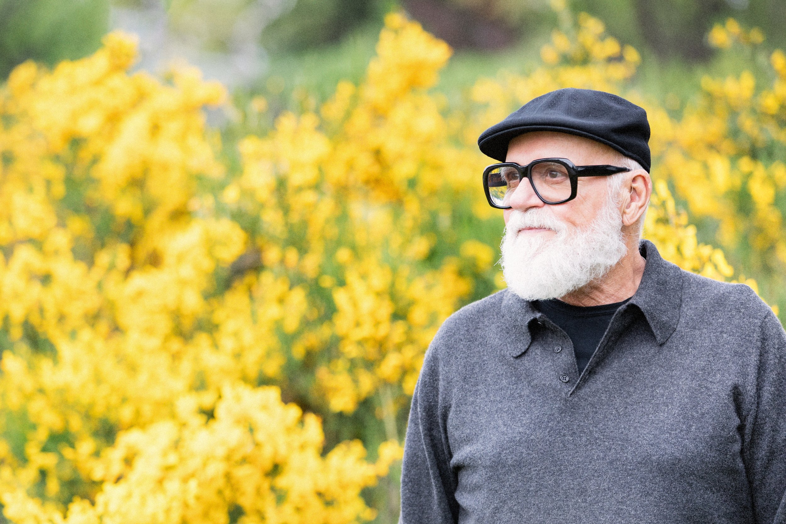 Older man white beard bold glasses posing outdoors on location wearing a hat and sweater.