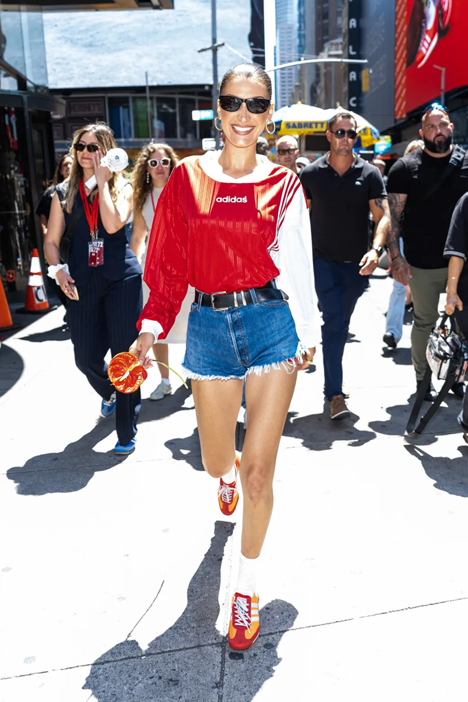 Model Bella Hadid wearing a red Adidas soccer jersey, denim cutoff shorts, black large sunglasses and red Adidas sneakers in Times Square.
