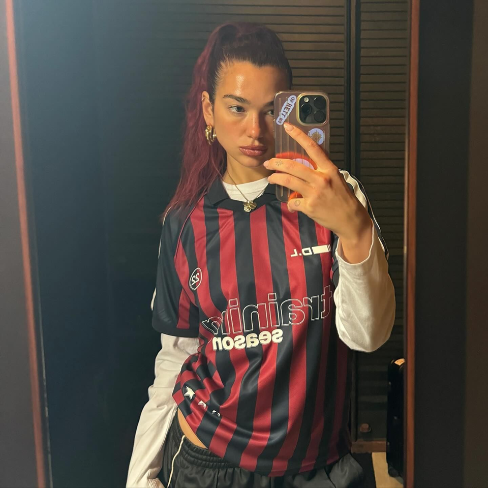 Artist Dua Lipa taking a mirror selfie wearing a soccer jersey and a long sleeve white shirt underneath it.