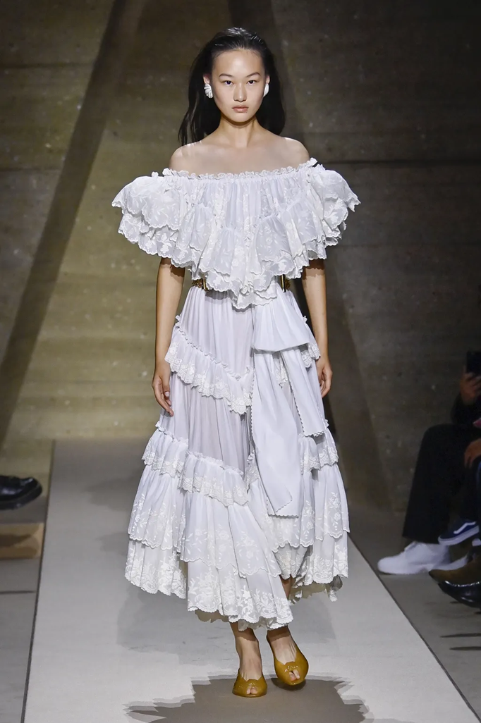 Image of an Asian model walking down the runway of a Chloe shoe wearing a white ruffled off the shoulder dress and camel colored jelly kitten heels.