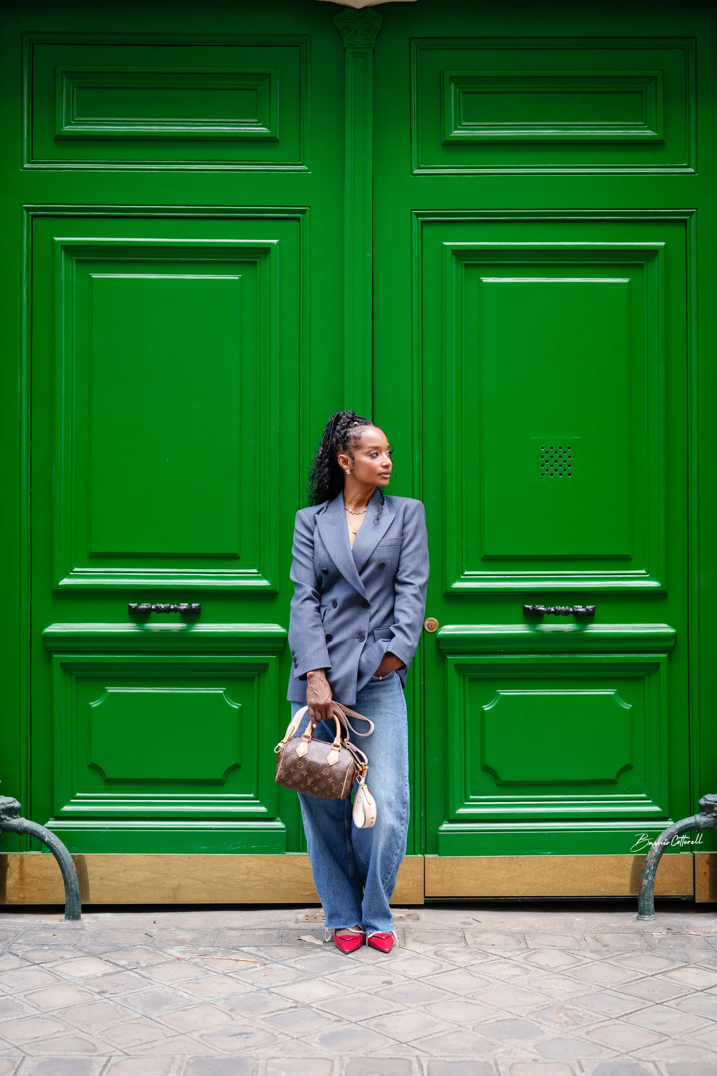 Person in an oversized double-breasted blazer, wide-leg jeans and red pointed flats, holding a brown monogram handbag in front of bright green doors