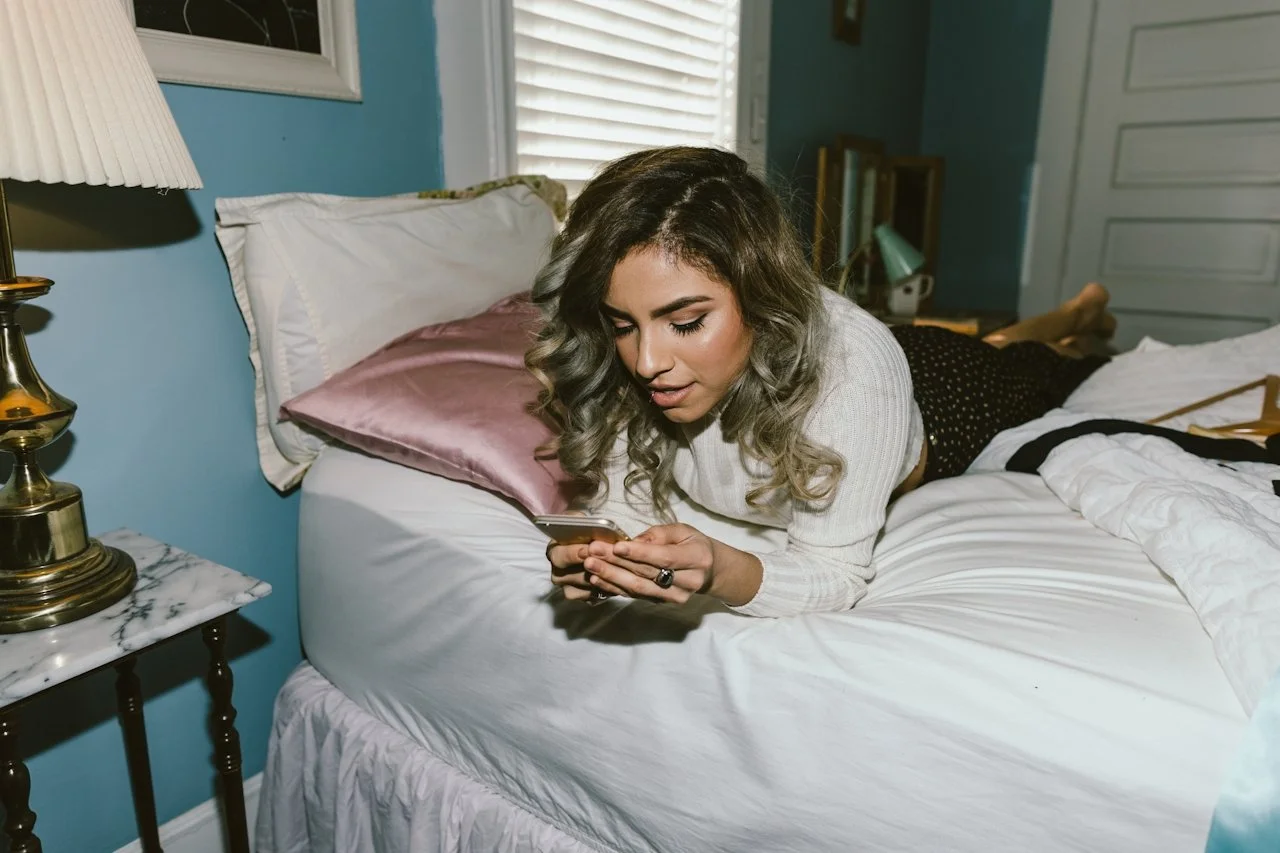 Image of a young woman lying on her bed on her stomach as she looks at a cell phone in her hands.