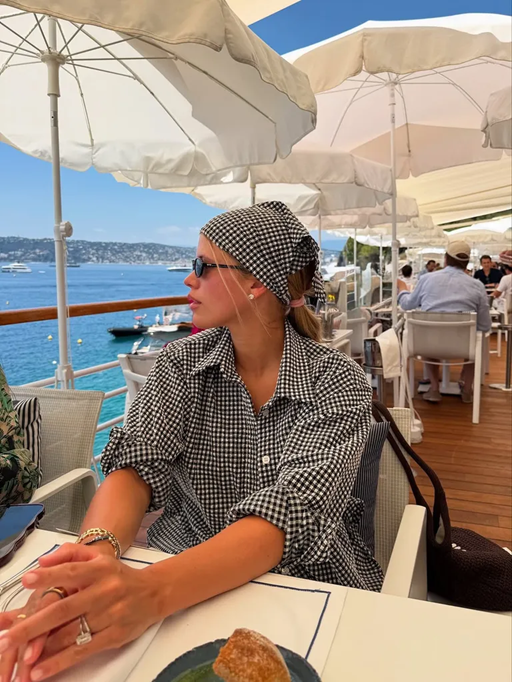Image of Sofia Grainge sitting at a sea-side restaurant looking out into the water wearing a gingham headscarf and matching shirt and small black sunglasses.