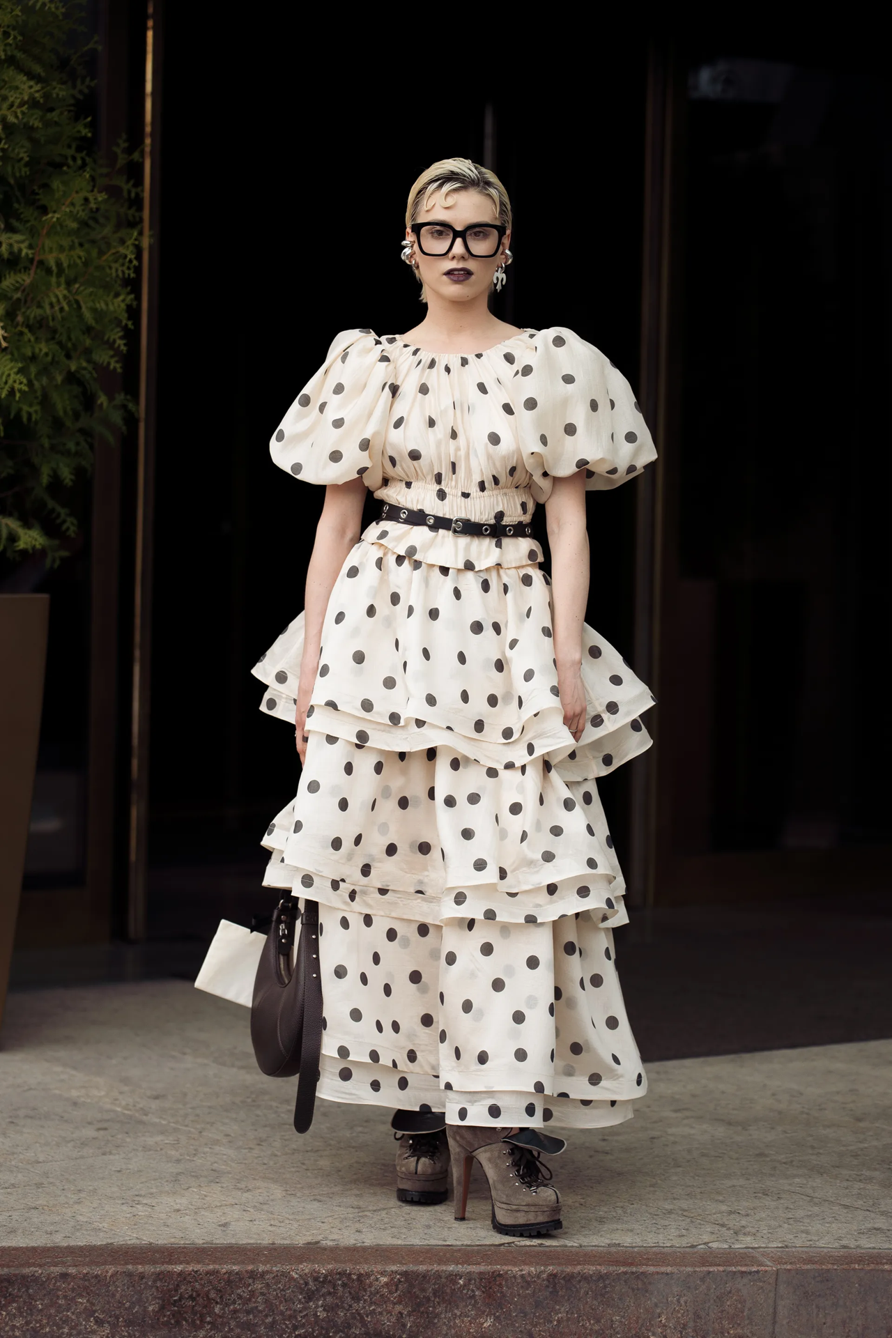 Photo of a Caucasian young woman with blonde hair and large square black frame glasses wearing a cream and black polka dot dress with boot high heels.