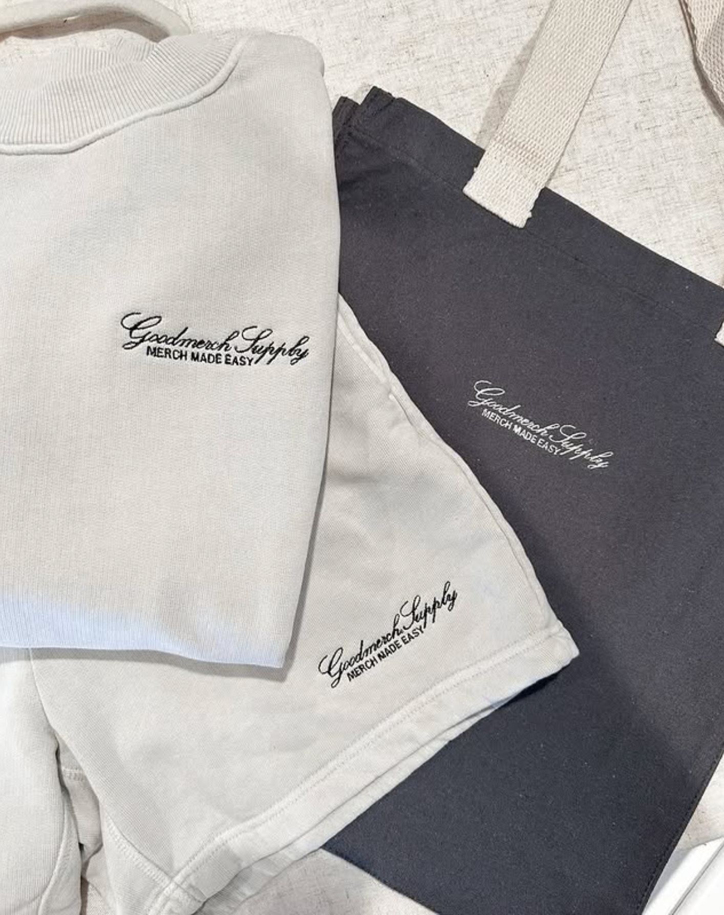 Multiple folded clothing items, including a white hoodie and dark pants, with embroidered text that says 'GoodMerch Supply MERCH MADE EASY' on the front of each piece.