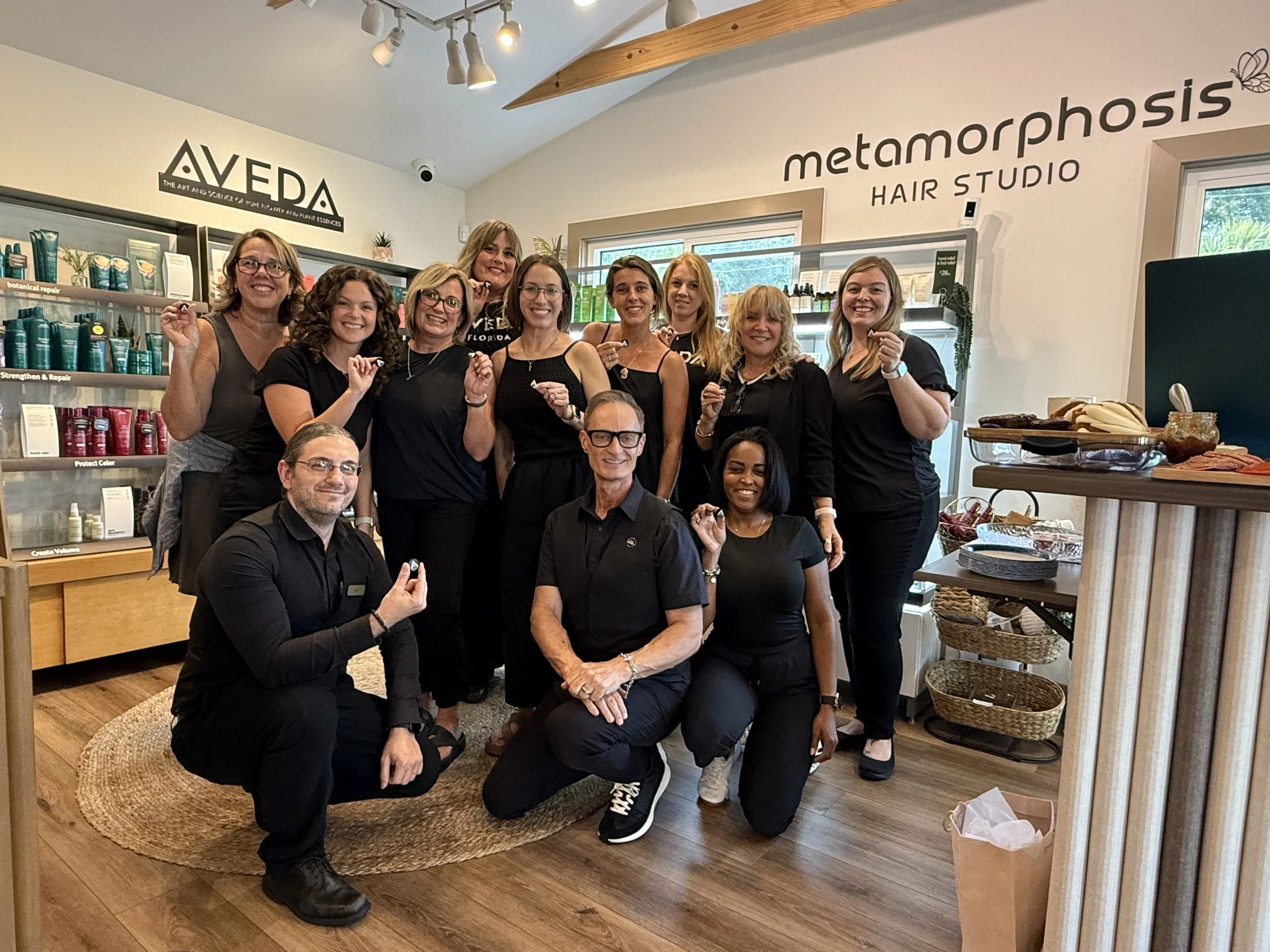 Group of ten people, some standing and some kneeling, posing inside a hair studio. The background features shelves with hair products and signage for Aveda and Metamorphosis Hair Studio. The group seems happy and is making small gestures with their hands.