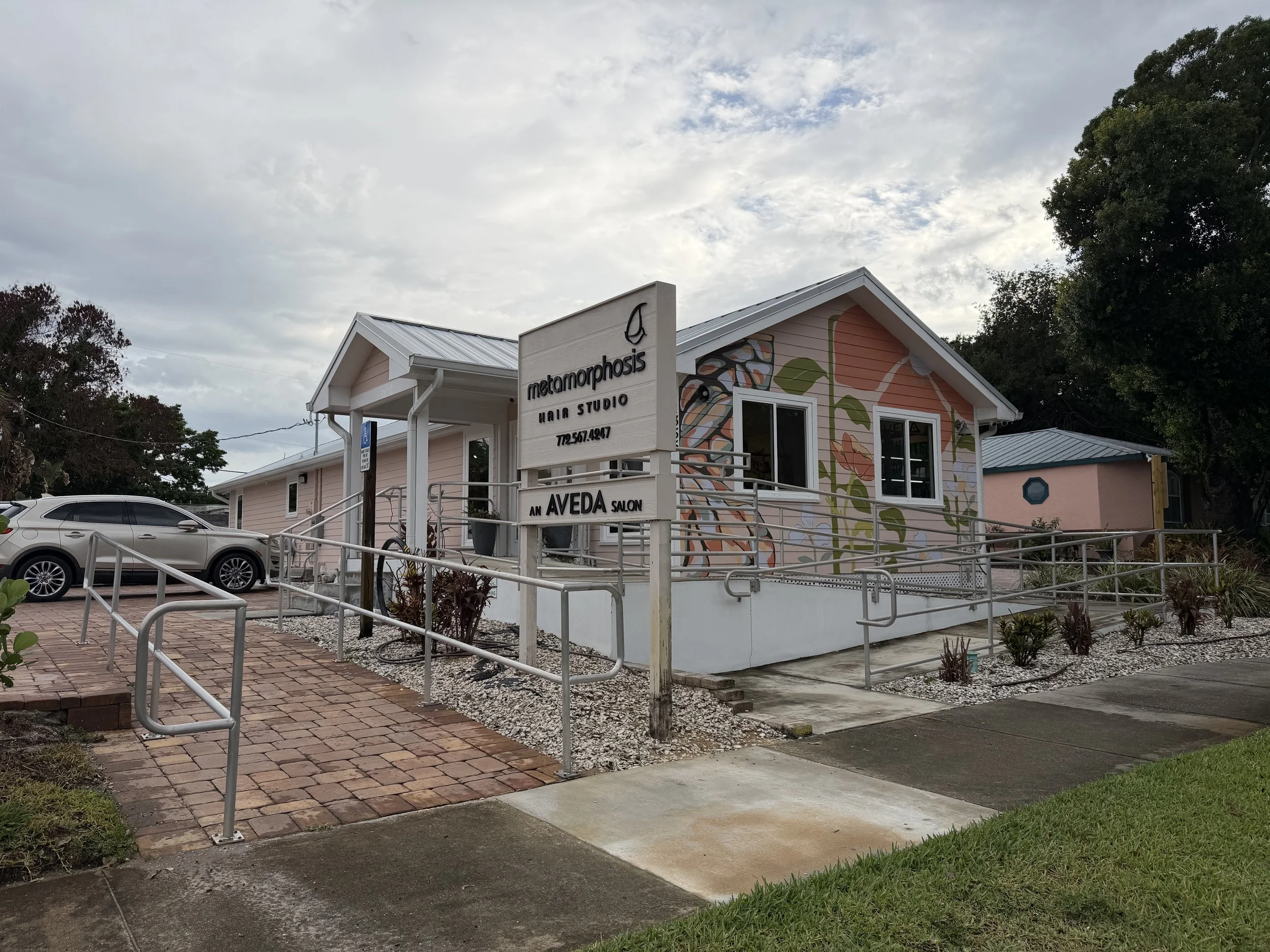 A pink building with a mural of flowers and leaves, with a sign that reads 'metamorphosis HAIR & SKIN STUDIOS' in front. There is a parking lot with cars and a wheelchair accessible ramp leading to the building. The sky is cloudy and trees are in the