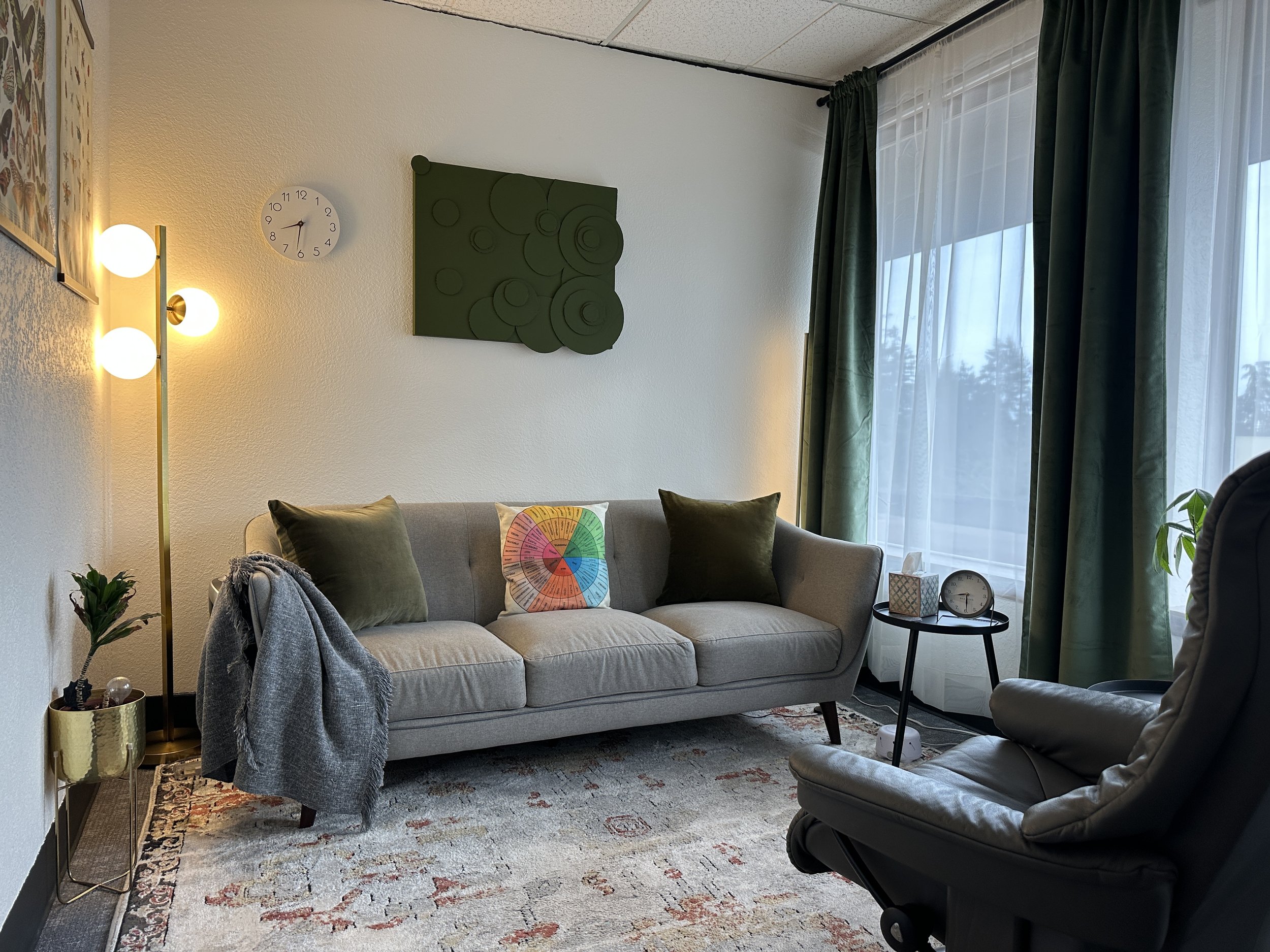 Cozy living room with a gray sofa, colorful pillows, and a gray blanket. A floor lamp with round bulbs is on the left, next to a small plant on a stand. A wall clock shows 10:12. Dark green curtains frame a large window, and a black side table holds 