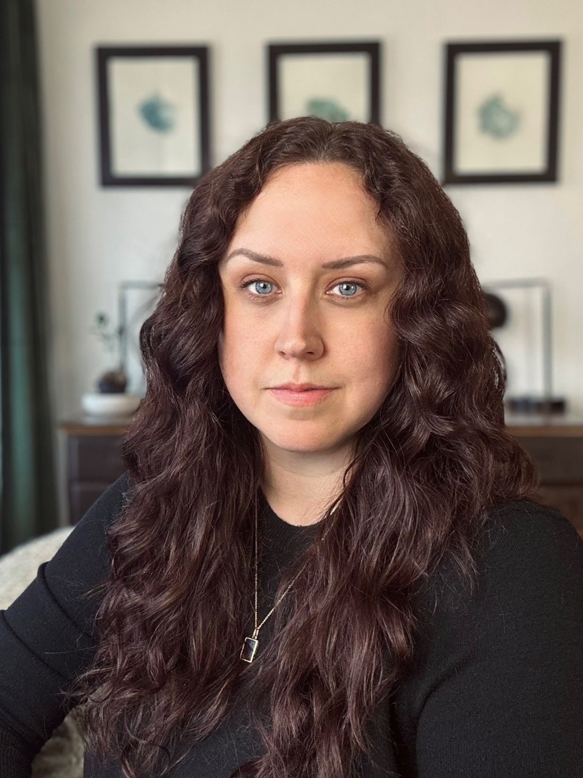 A woman with long curly hair wearing a black top and a necklace, sitting indoors with a neutral expression. There are framed pictures on the wall in the background.