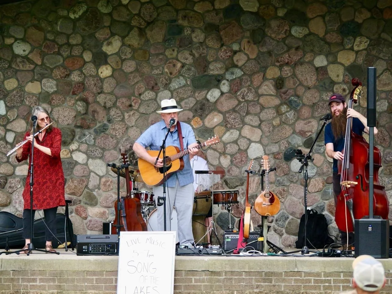 A band performing live music outdoors on a stage with a stone wall background. The band members include a woman playing a flute, a man singing and playing an acoustic guitar, and a man with a beard playing an upright bass. There are multiple guitars 