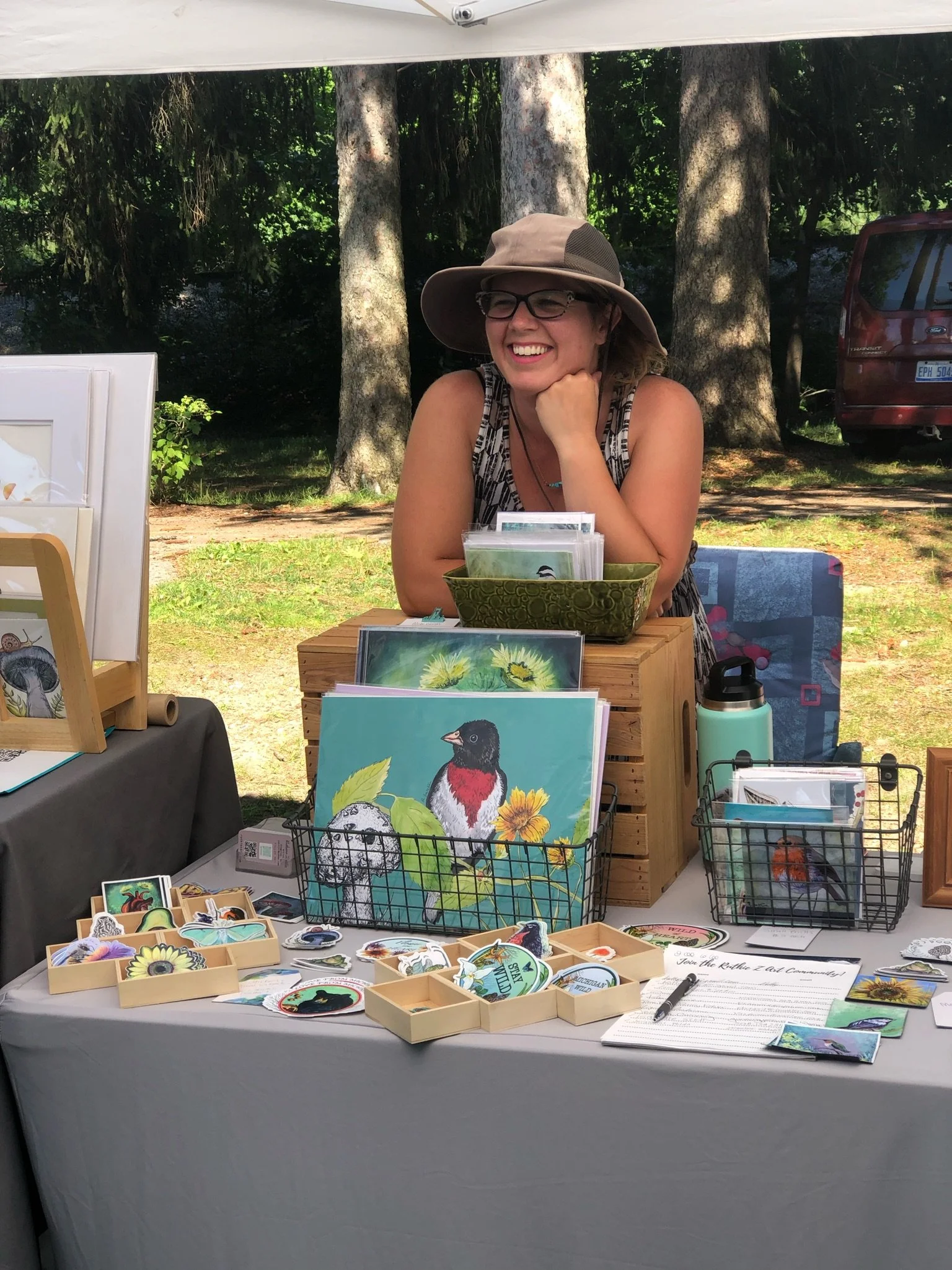 A smiling woman wearing a wide-brimmed hat and glasses sits behind a display of colorful artwork and stickers outdoors, with tall trees and a red vehicle in the background.