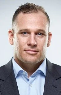 Professional headshot of a man in a suit and light blue shirt, against a plain background.