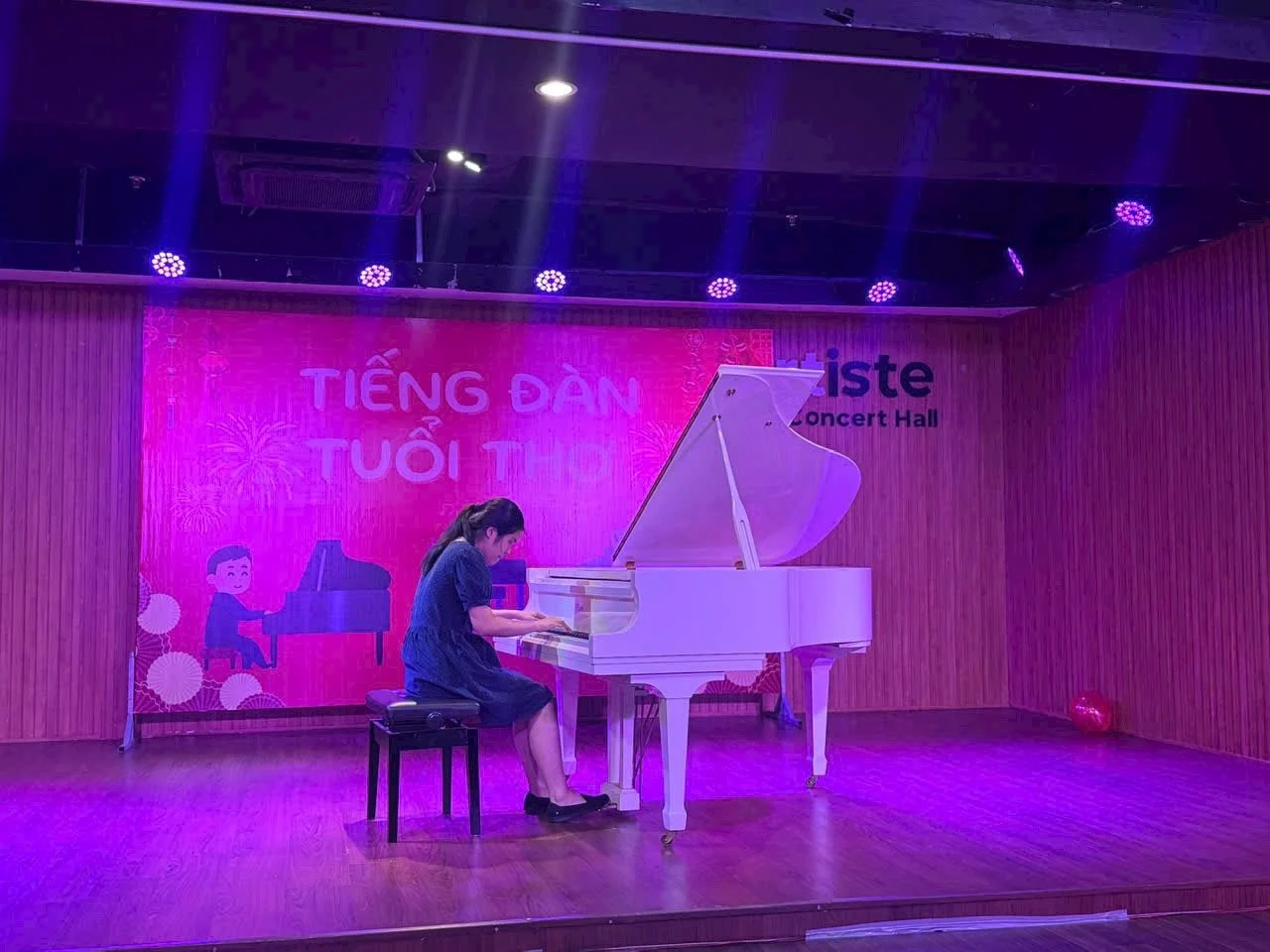 A girl playing a white grand piano on a stage with wooden walls and purple lighting. A large pink banner with Vietnamese text and illustrations of fireworks and children is in the background.