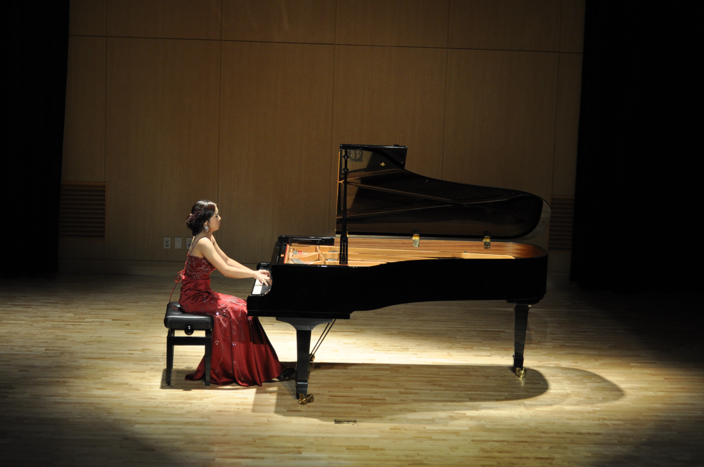 A woman in a red gown playing a grand piano on a stage with a spotlight.