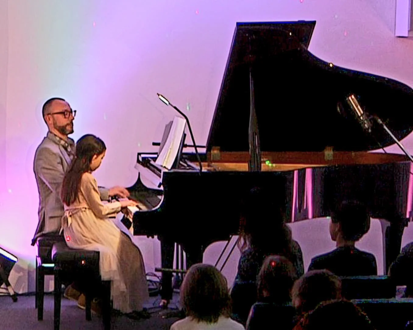 A man and a young girl playing a grand piano on stage with an audience watching.