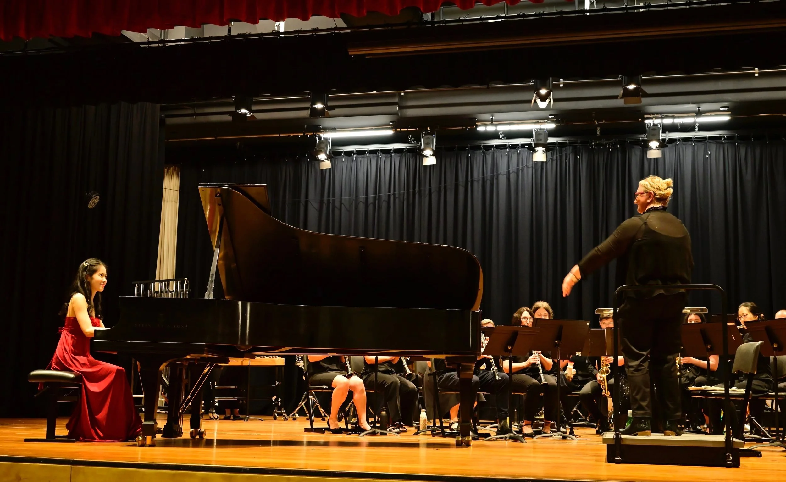 A young girl in a red dress sitting at a grand piano on stage during a performance. A woman appears to be conducting the orchestra, with other musicians seated behind. The stage has a black curtain backdrop and stage lights hanging from the ceiling.
