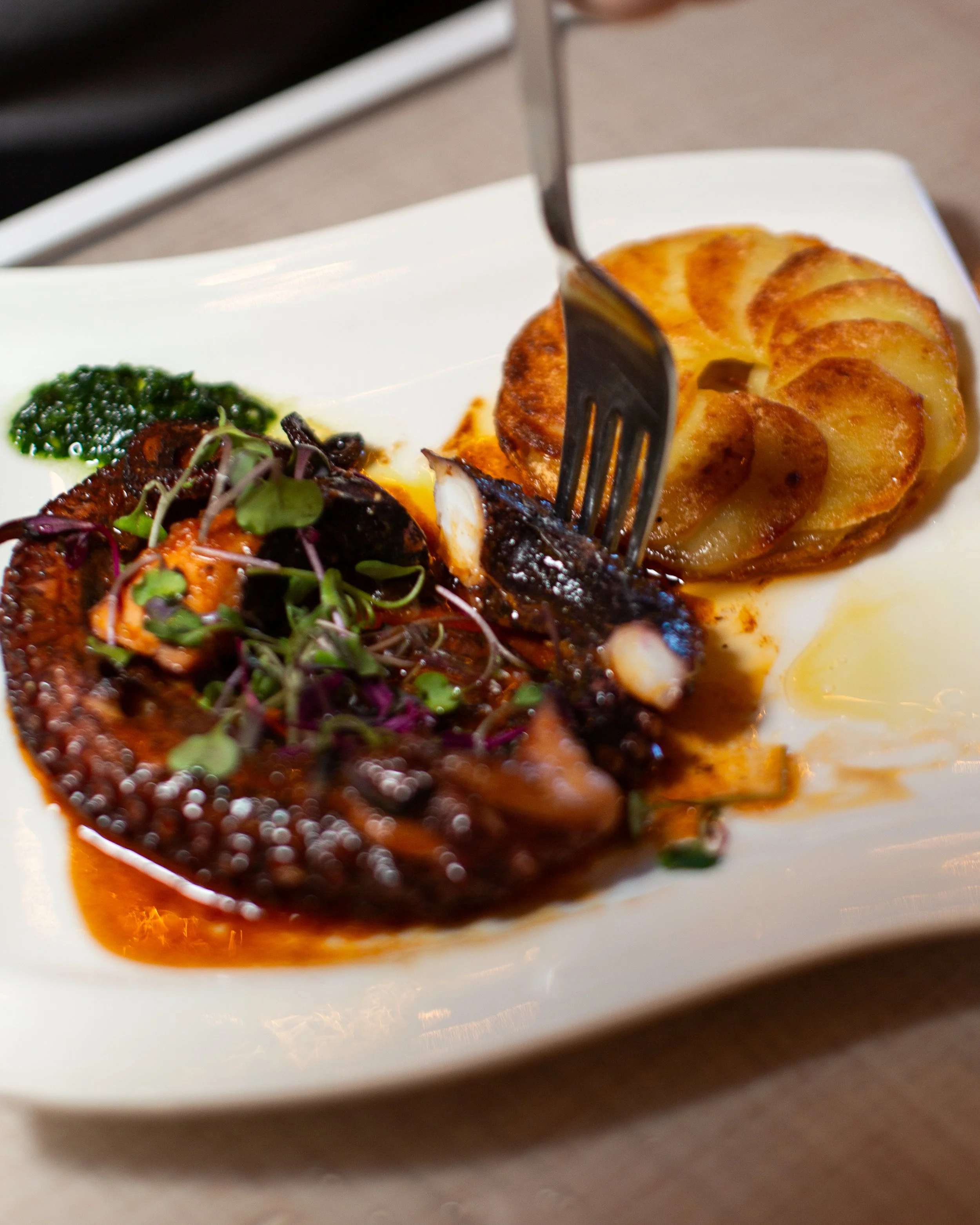 Close-up of a plated dish featuring roasted potatoes with a golden-brown surface, a serving of green pesto, and a piece of grilled meat topped with microgreens, on a white plate.