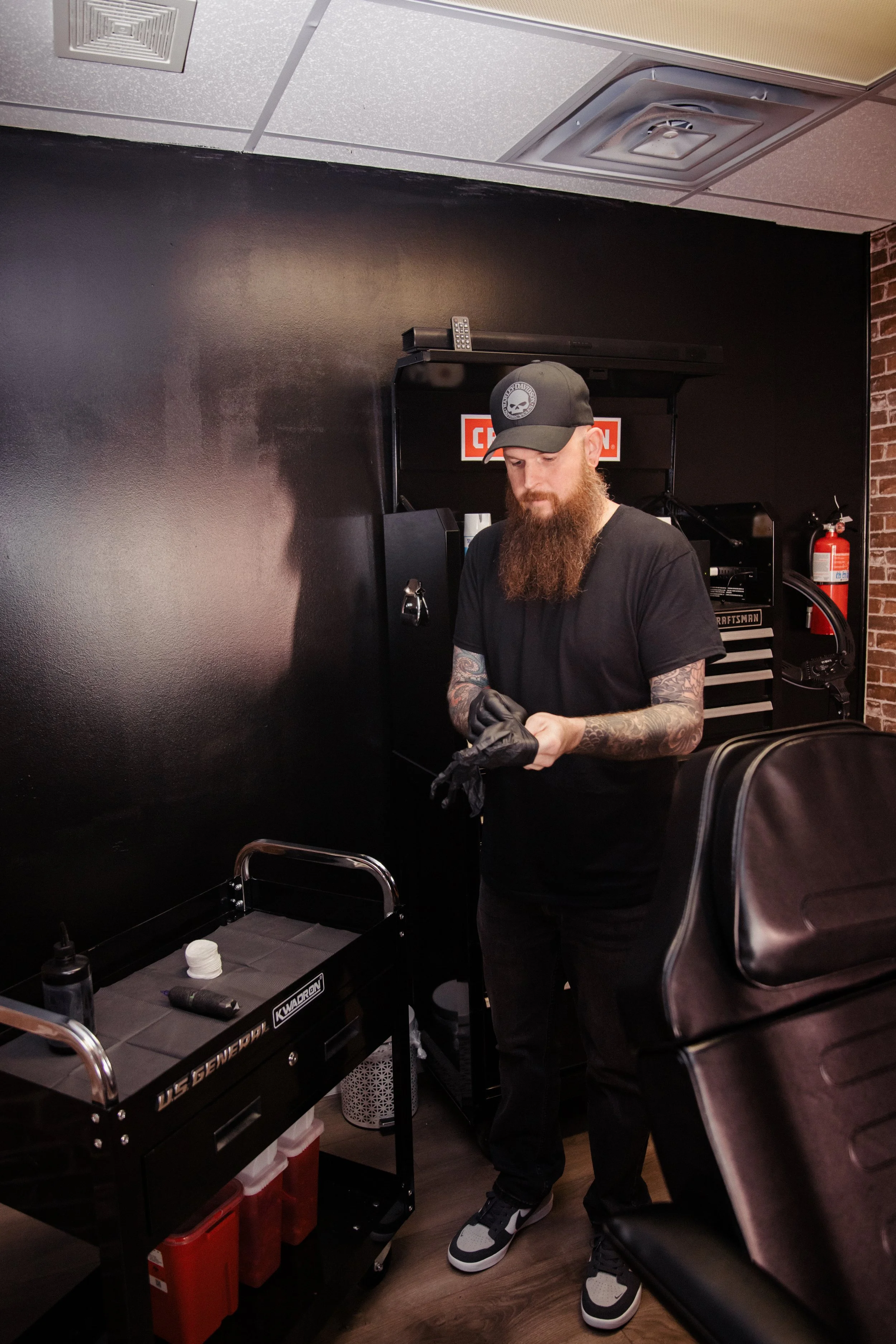 A man with a beard and tattoos, wearing a black cap and black t-shirt, putting on black gloves in a tattoo studio. There is a tattoo cart with supplies and a black tattoo machine behind him.
