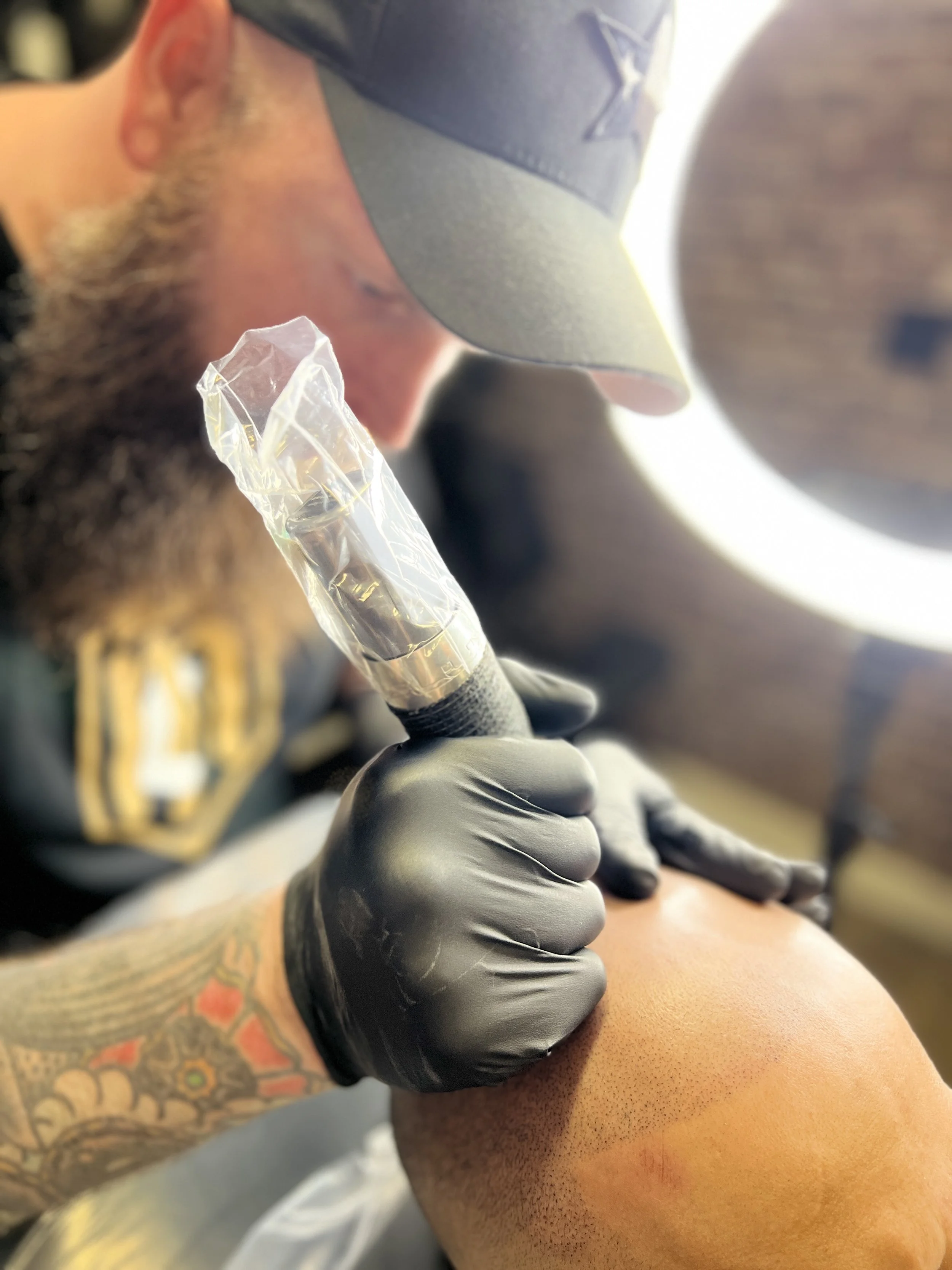 Tattoo artist wearing black gloves using a tattoo machine on a person's arm in a tattoo shop.