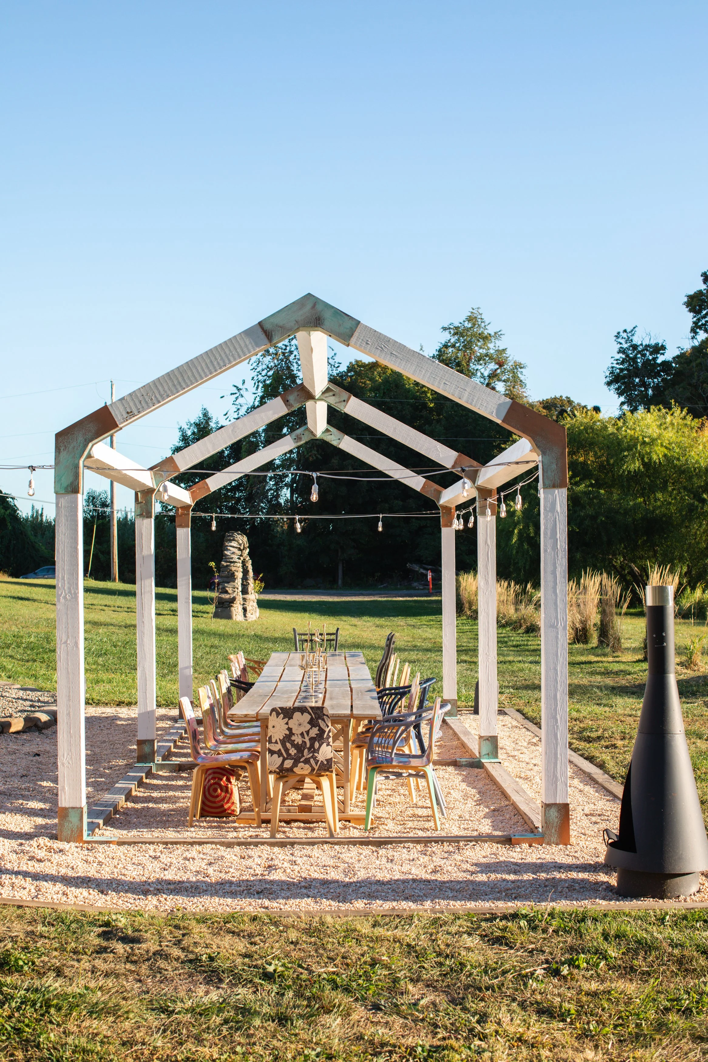 Outdoor dining area under a wooden pergola with string lights, surrounded by grass and trees.