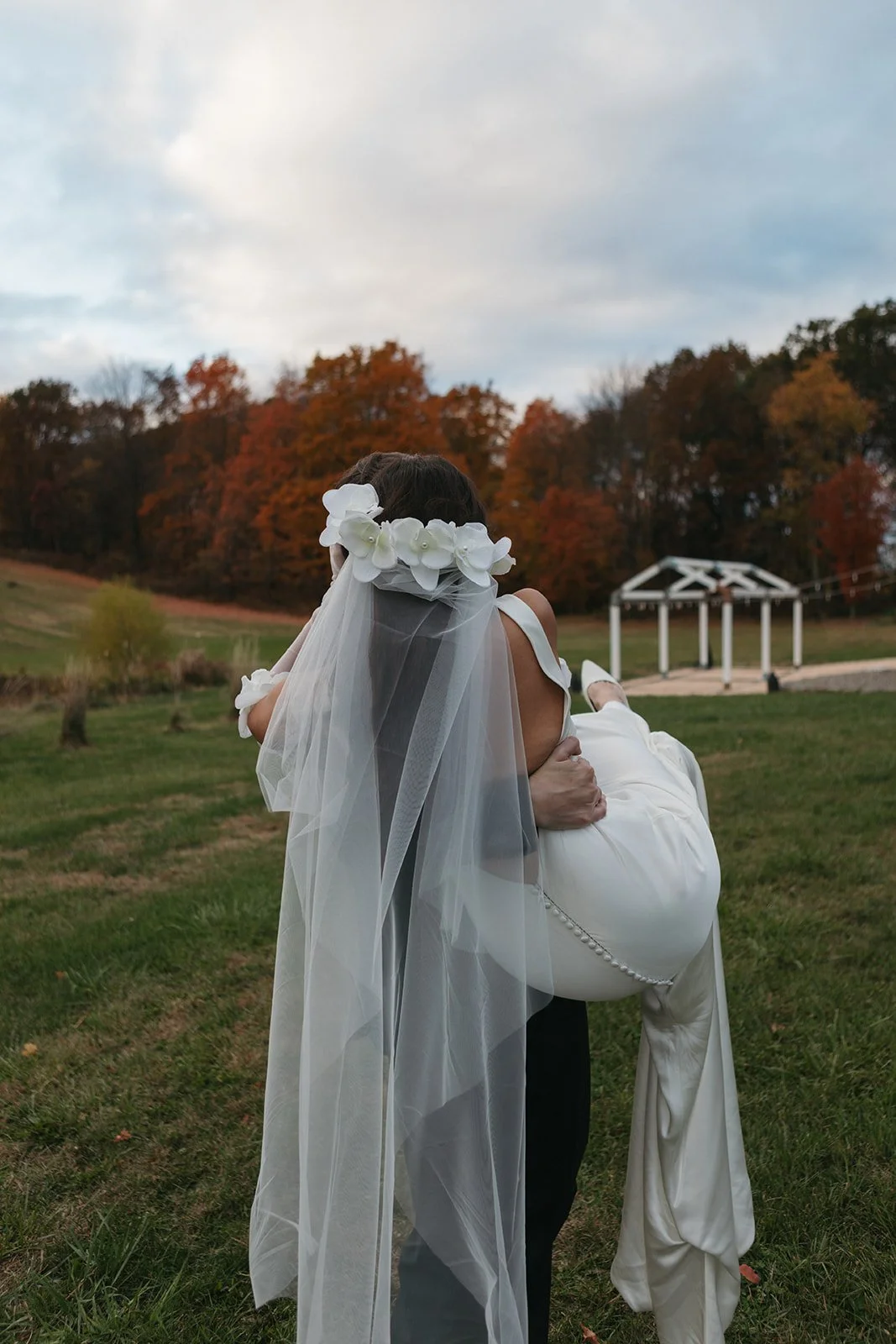 A person dressed in wedding attire, carrying a bride in a white gown and veil with white flowers, outdoors with fall foliage in the background.