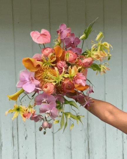 Hand holding a colorful bouquet of mixed flowers with pink, yellow, orange, and purple blooms in front of a light blue wooden background.