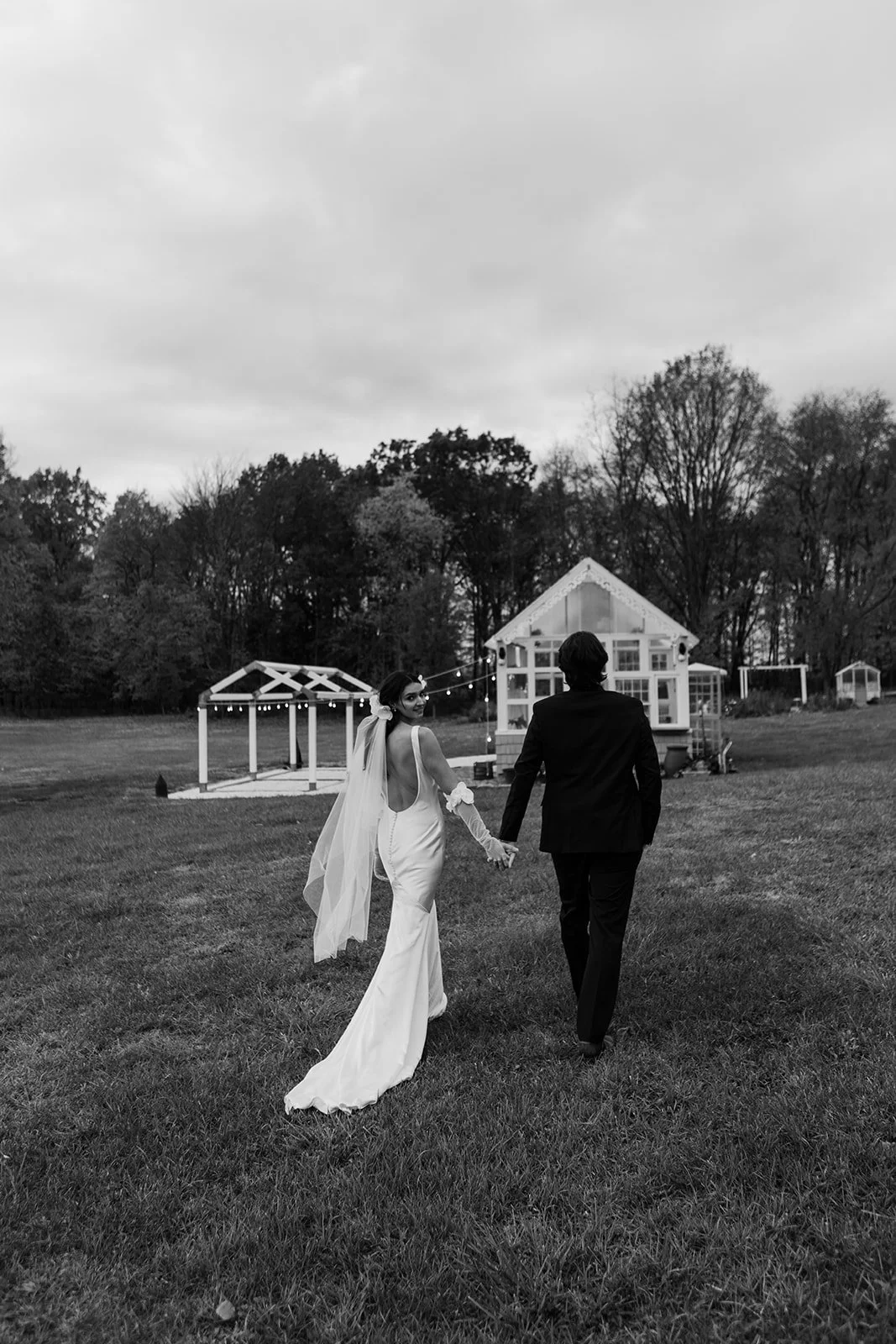 A couple in wedding attire holding hands, walking away from the camera on a grassy field, with a small house and trees in the background, in black and white.
