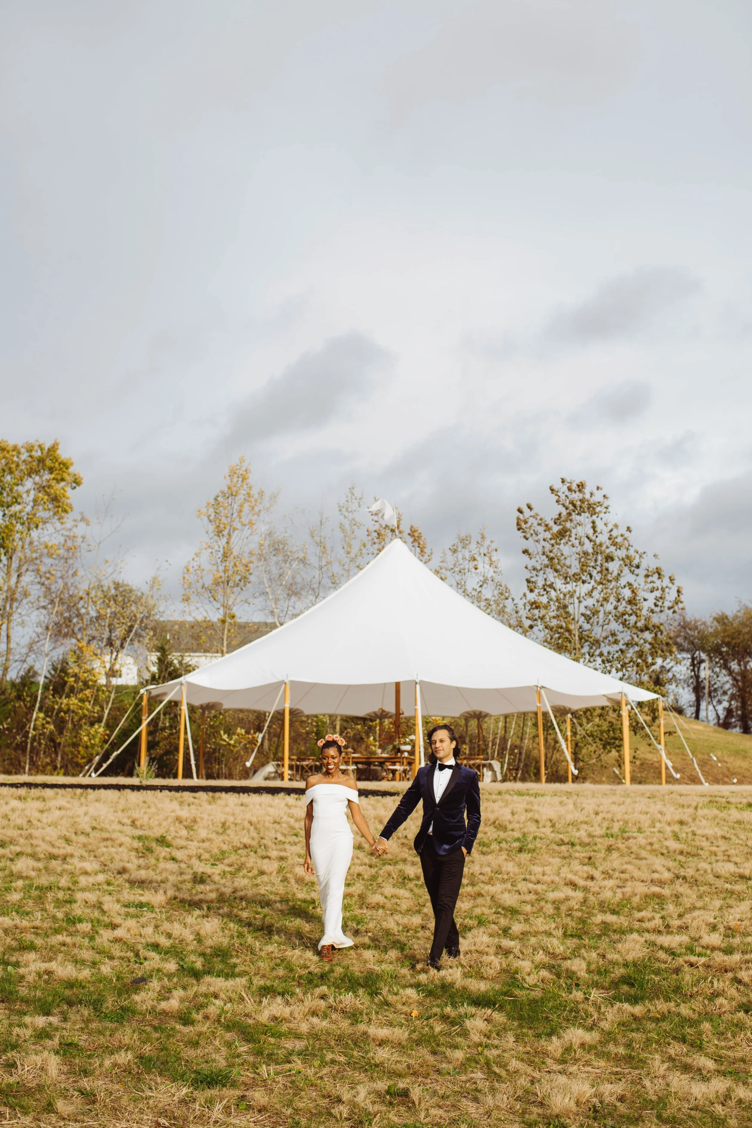 A couple walking hand in hand on a grassy field in front of a large white event tent during daytime. The woman is in a white dress, and the man is in a dark suit with a bow tie. The sky is overcast, and trees are visible in the background.